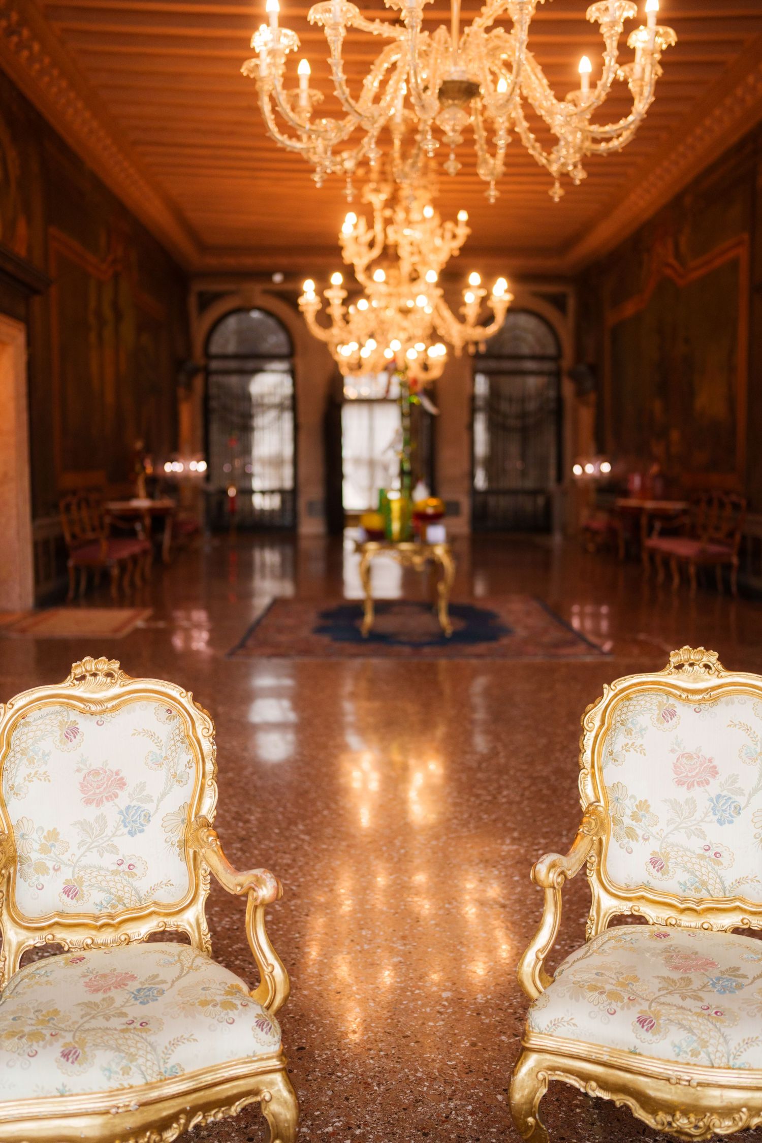 Elegant baroque chairs and crystal chandeliers adorn a luxurious palace hallway with polished marble floors.