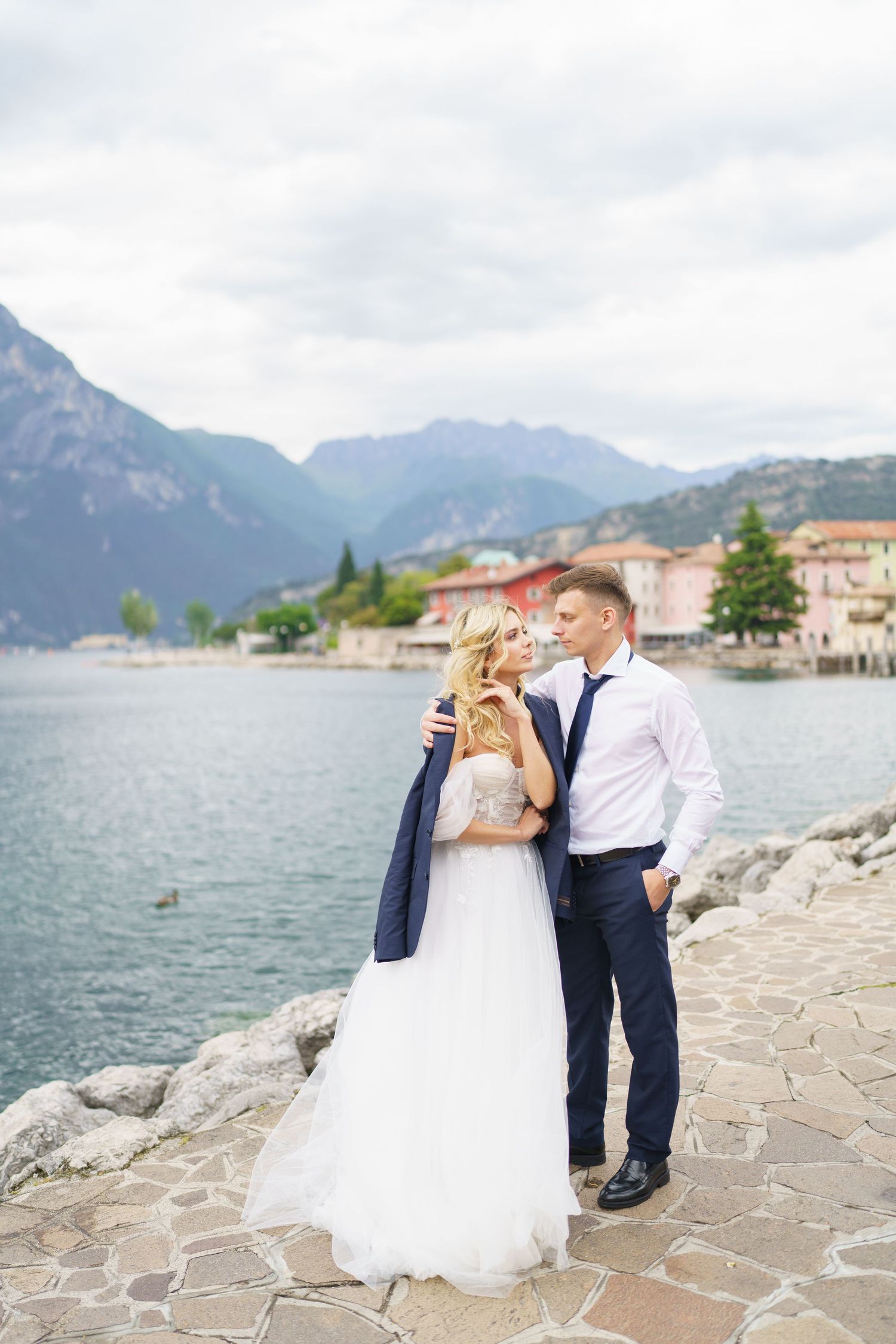 A couple shares a romantic moment on a stone walkway overlooking a scenic mountain lake with buildings in the background.