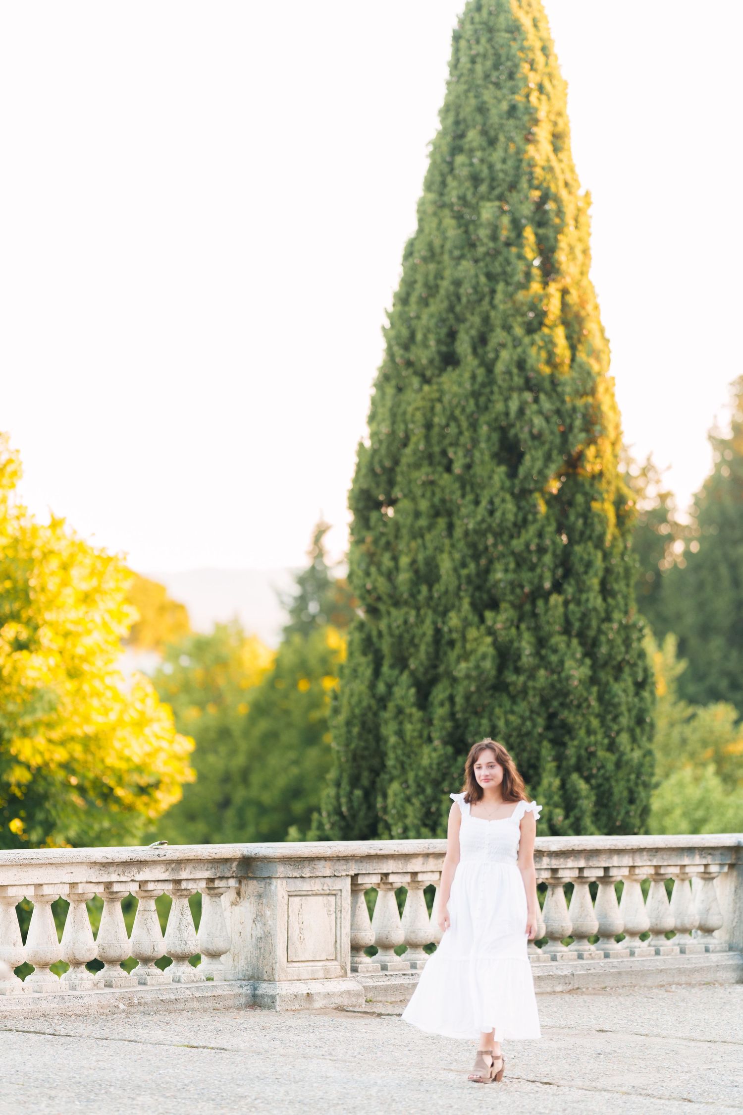 A person in a white dress stands by an ornate stone balustrade in a garden at golden hour.