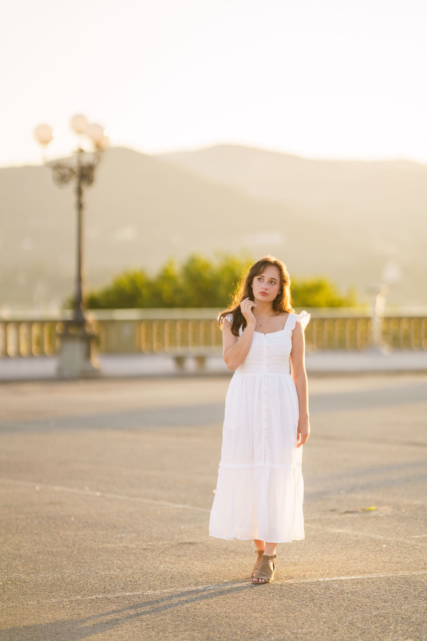 A small figure in a white dress walks across a sunlit concrete surface with mountains in the background at golden hour.