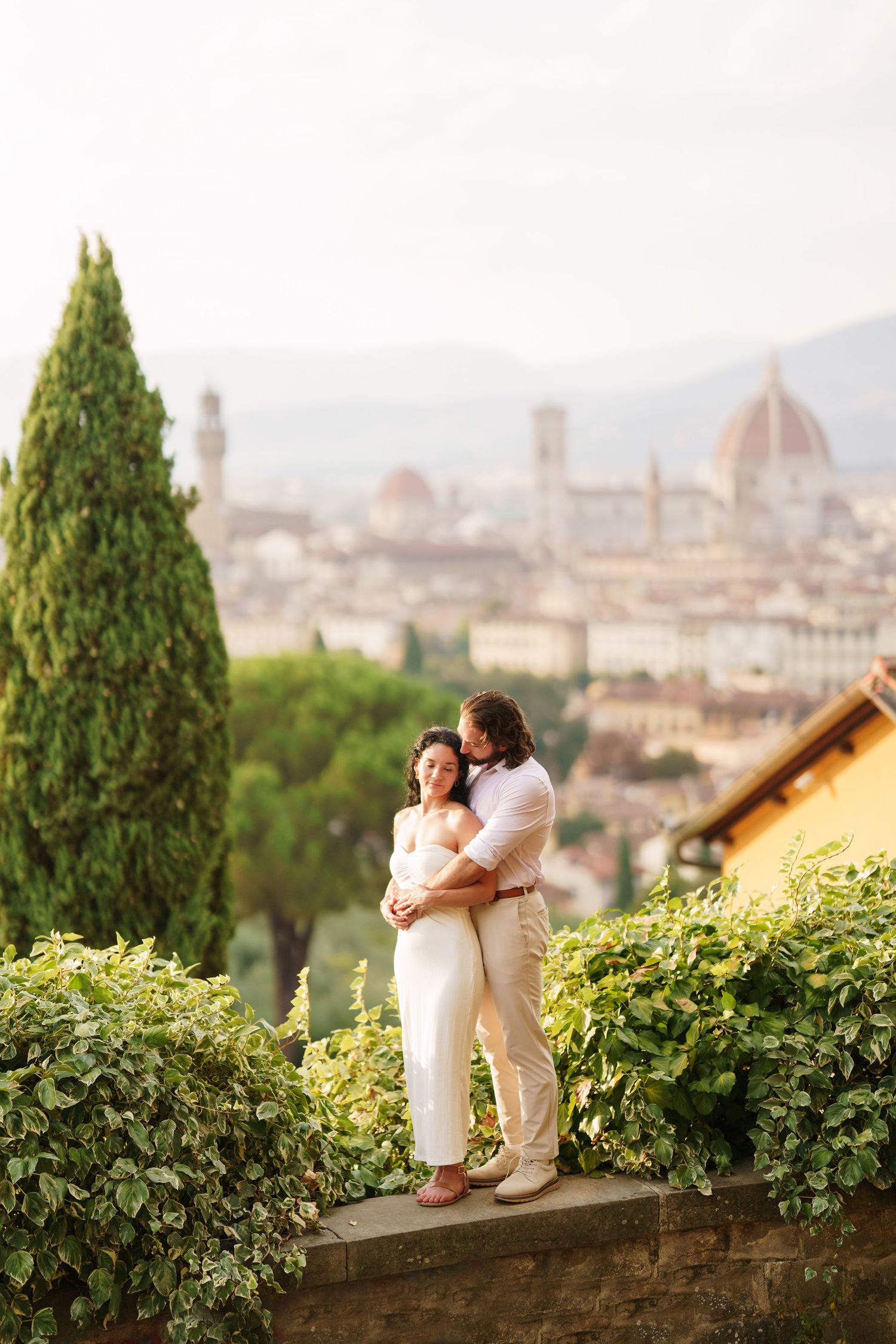 Couple in white outfits embrace while sitting on stone wall overlooking Florence cityscape with cypress trees and greenery.