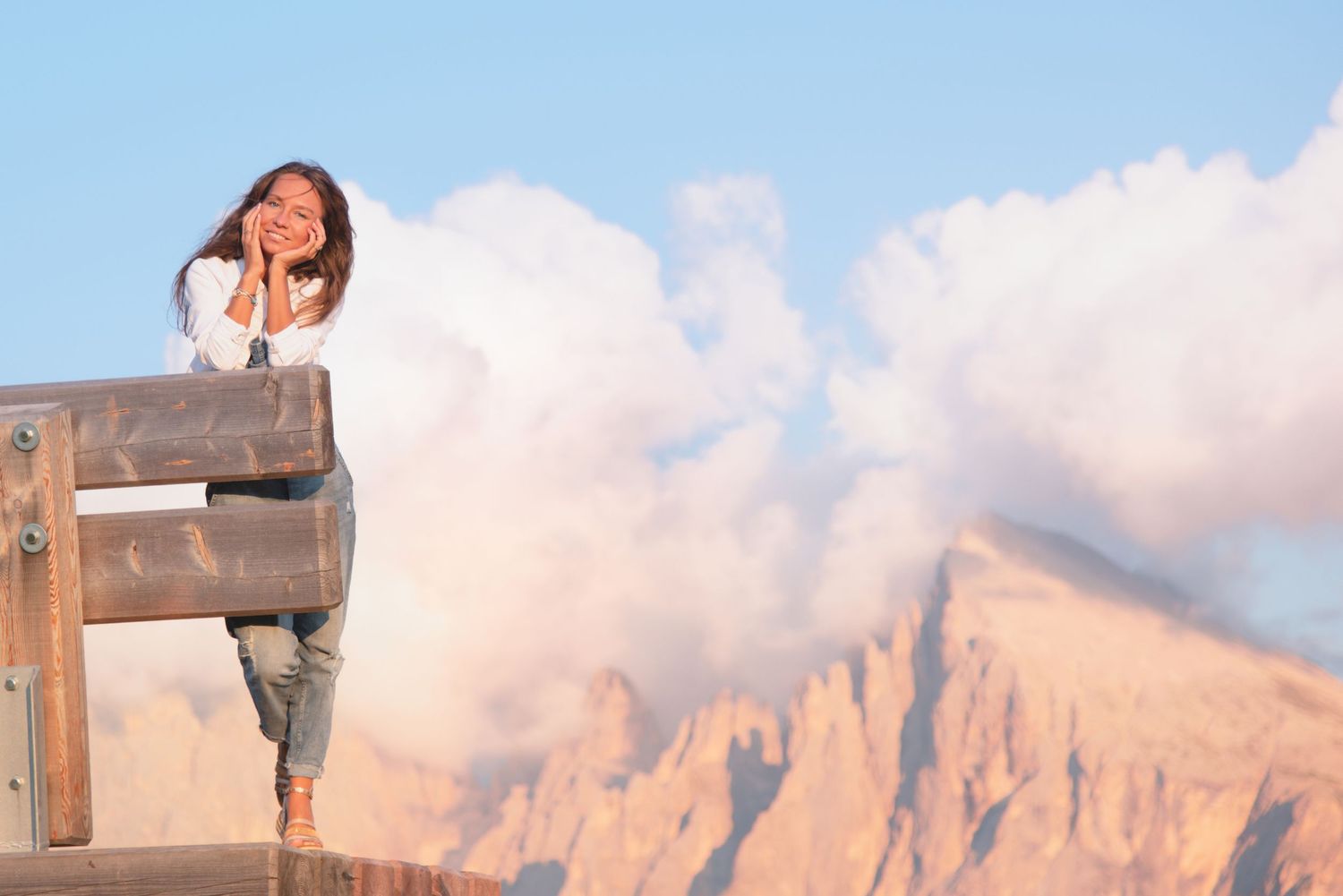 Person sitting on wooden railing at scenic mountain viewpoint with cloudy sky and rocky peaks in background.
