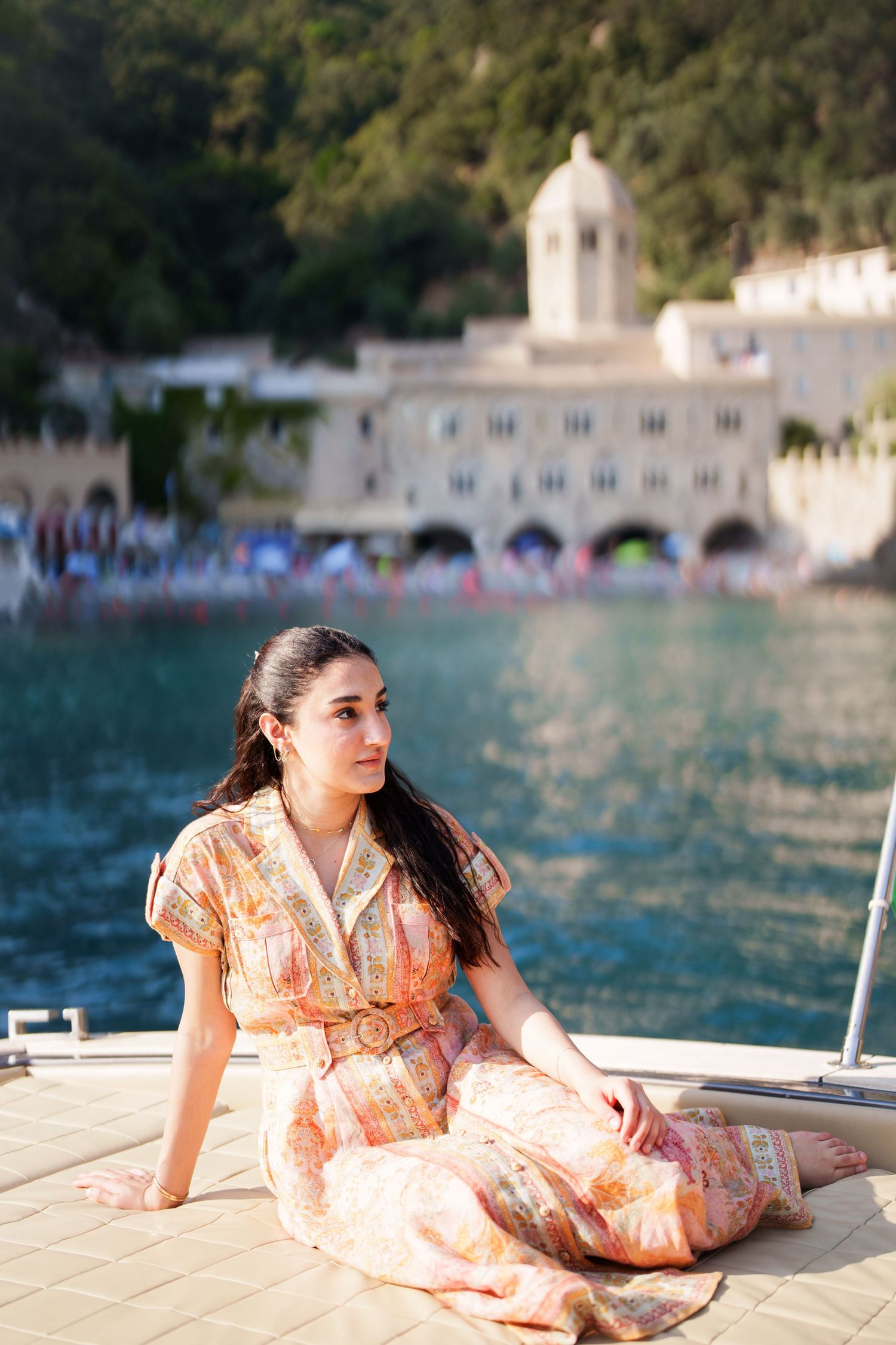 Young woman in floral maxi dress sitting on yacht deck with Mediterranean village and turquoise water behind her.