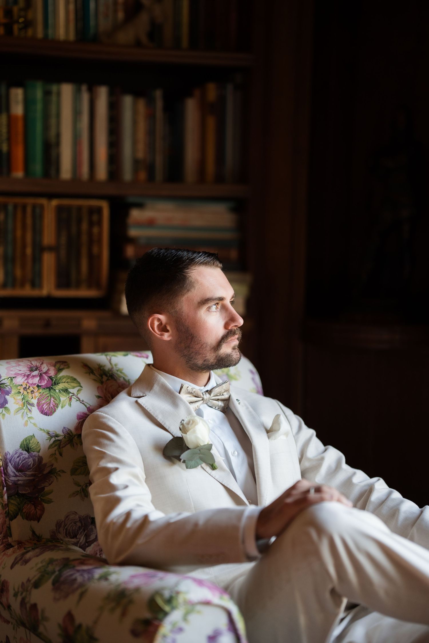 Elegant groom in white suit with floral boutonniere sits in a vintage armchair before a wooden bookshelf.