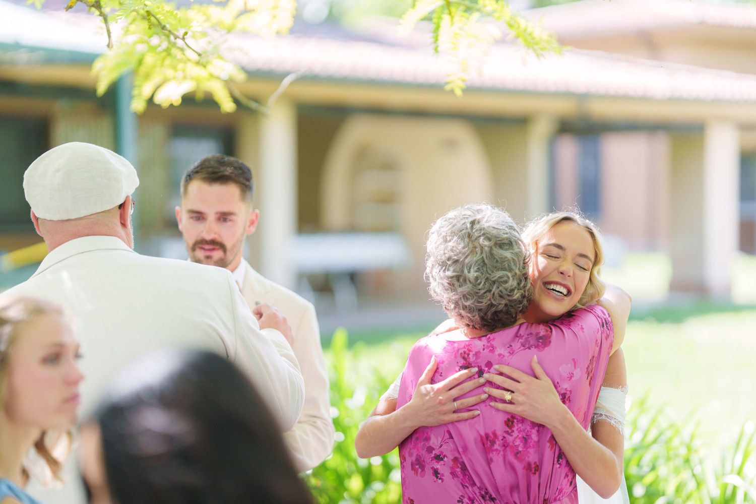People hugging and socializing outdoors at a sunny garden gathering near a house.