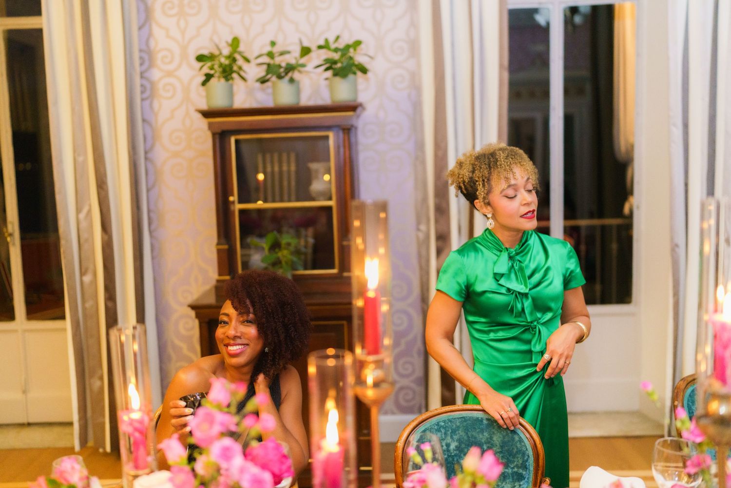 Two women in elegant green dresses laugh at a candlelit dinner table adorned with pink floral arrangements.