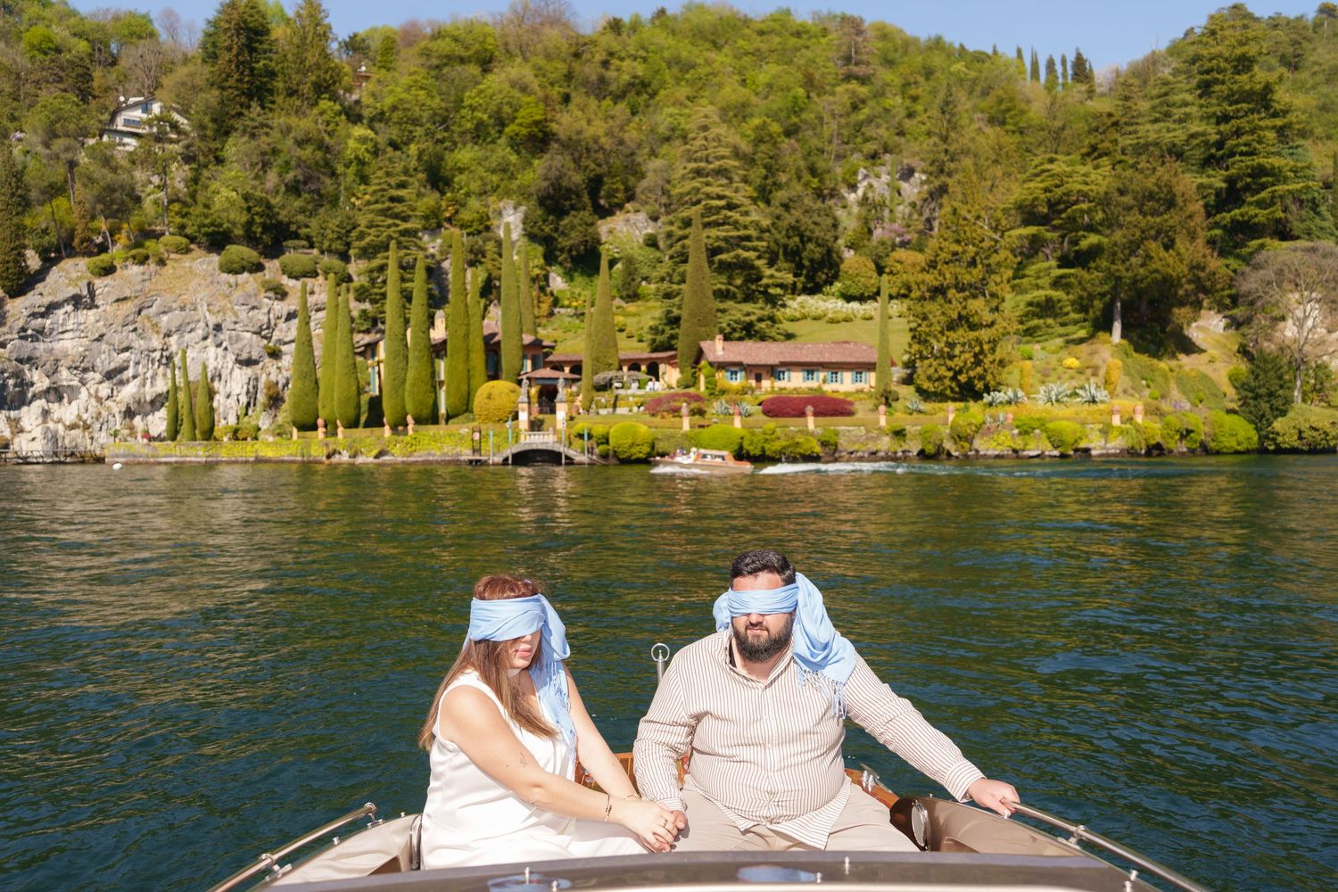 Couple in blue headbands riding a wooden boat on a lake with lush green hills and a villa in the background.