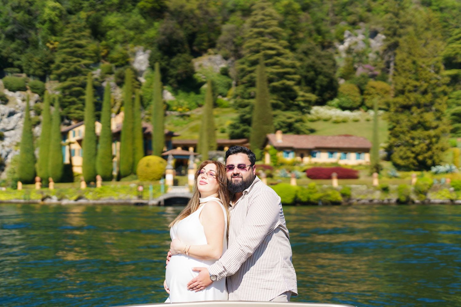 Couple embracing on a boat with lush greenery and a villa on Lake Como in the background.