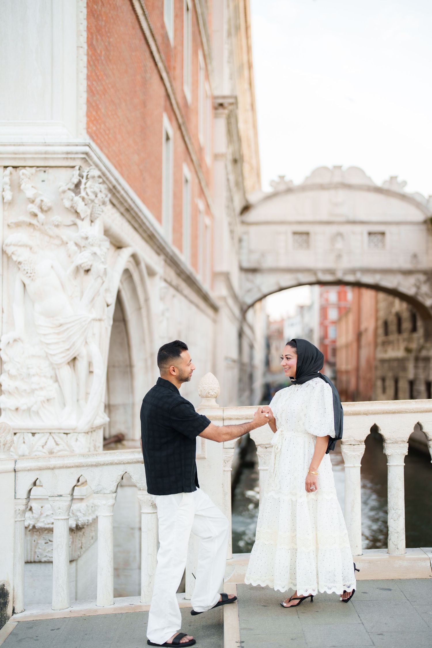 Couple holding hands near the Bridge of Sighs in Venice, woman in white dress and hijab, man in black shirt.