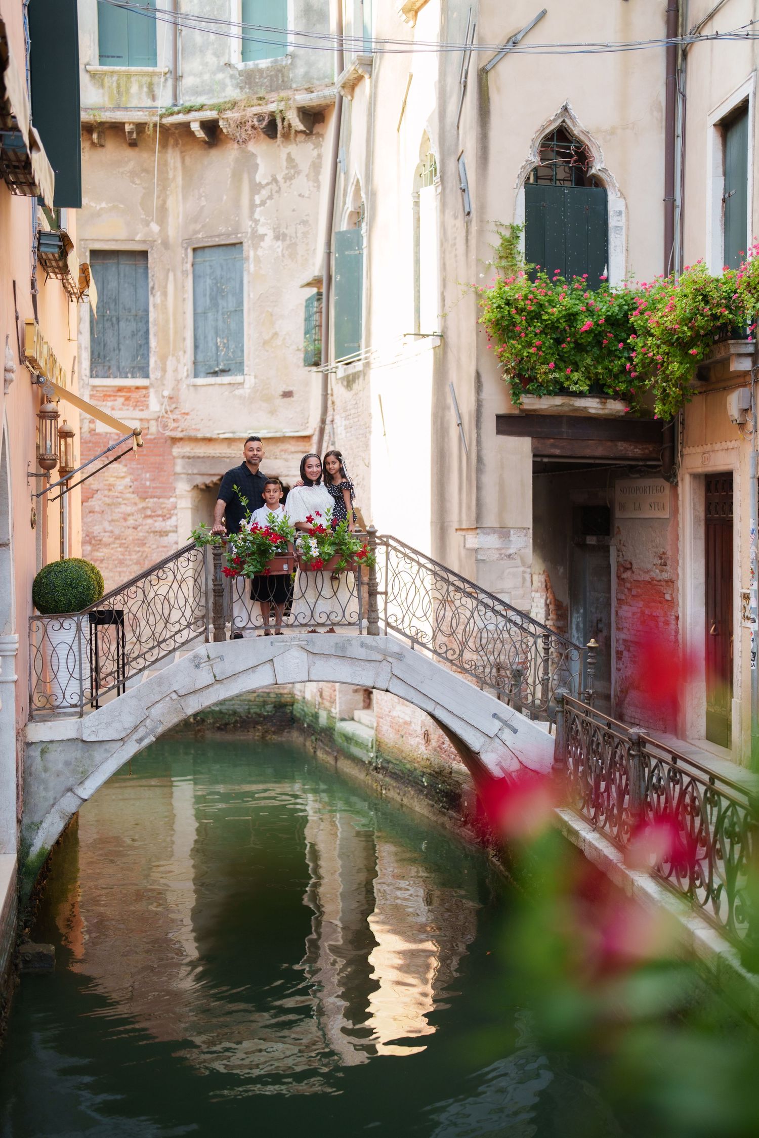 Family posing on a charming stone bridge over a Venice canal, surrounded by colorful flowers and historic buildings.