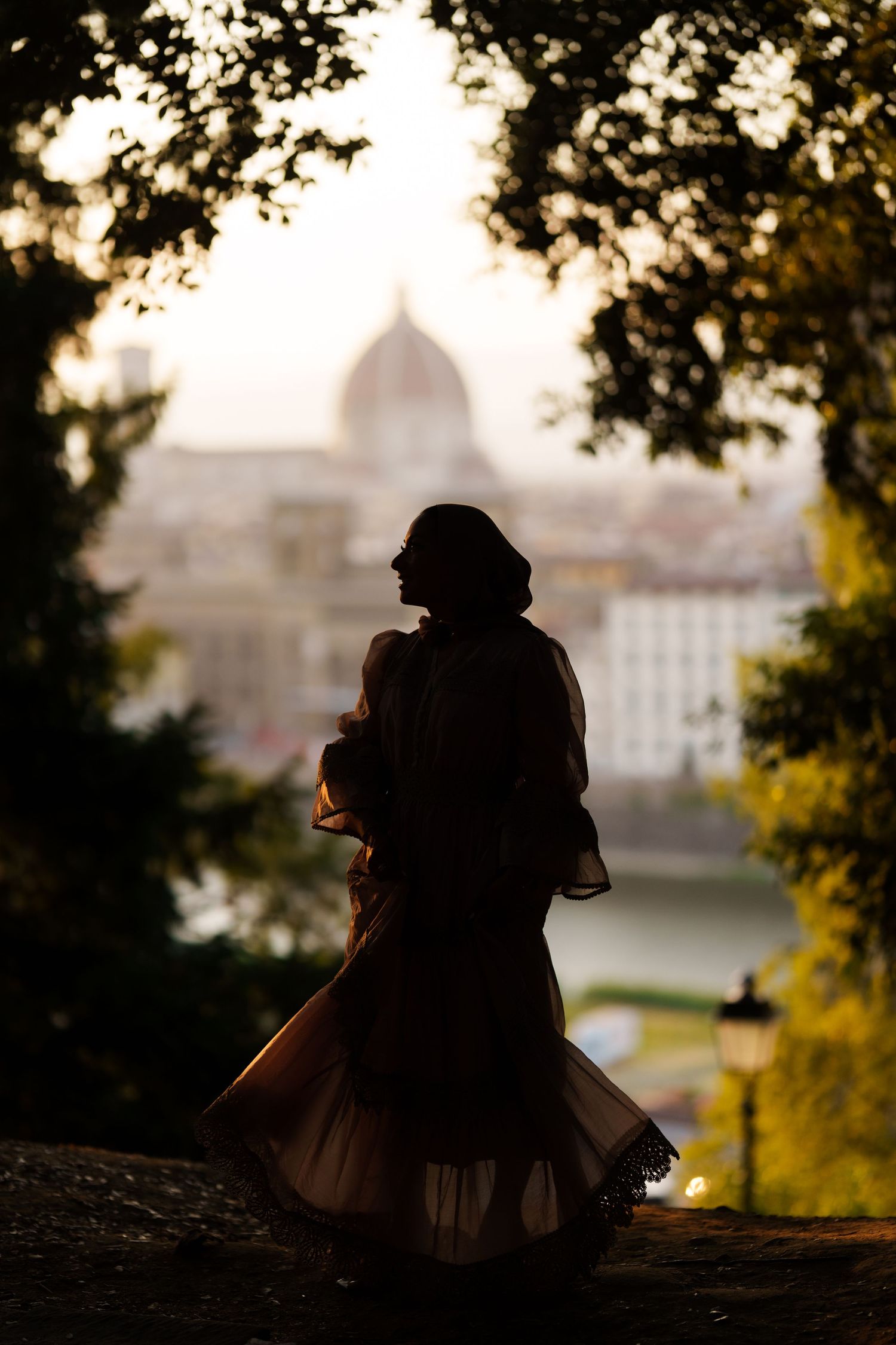 Silhouette of a woman in flowing dress overlooking Florence skyline with cathedral dome at golden hour.