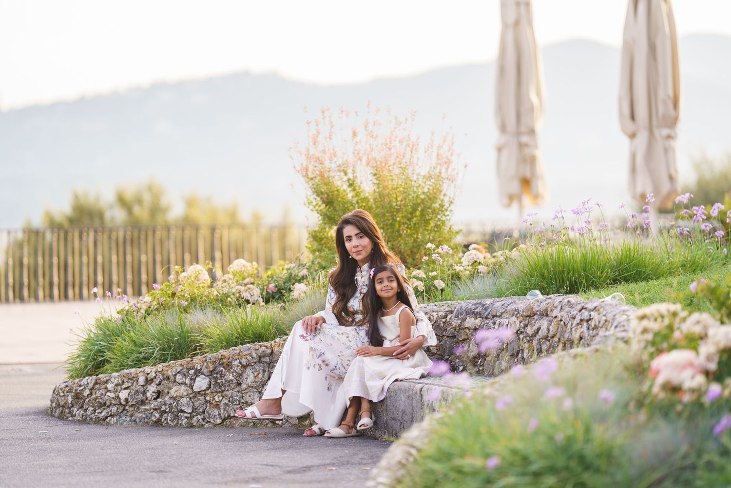 A mother and daughter in matching white dresses sit on stone steps surrounded by blooming flowers with mountain views.