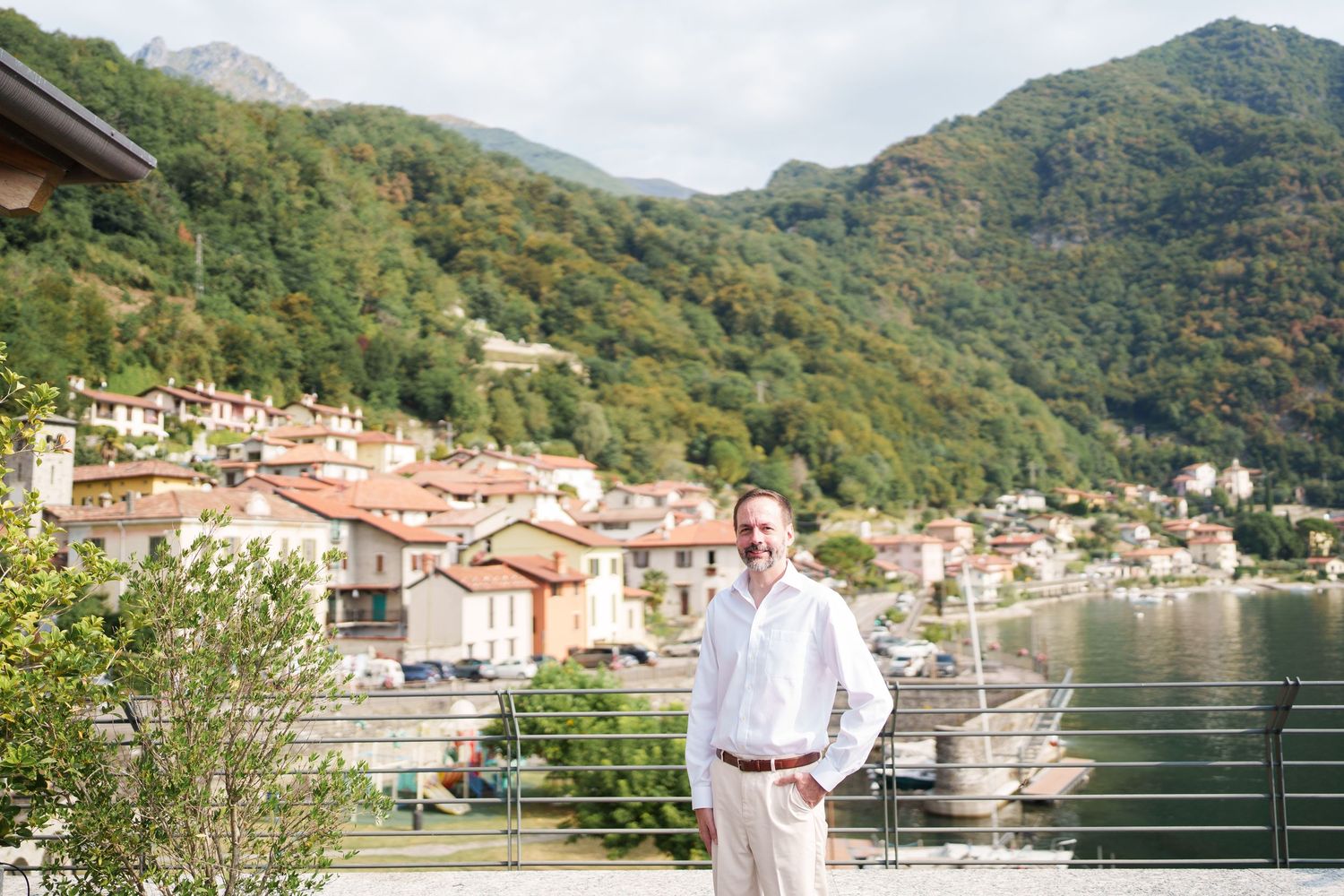 Man in white shirt smiling by Lake Como, Italy, with colorful village and green hills in background.