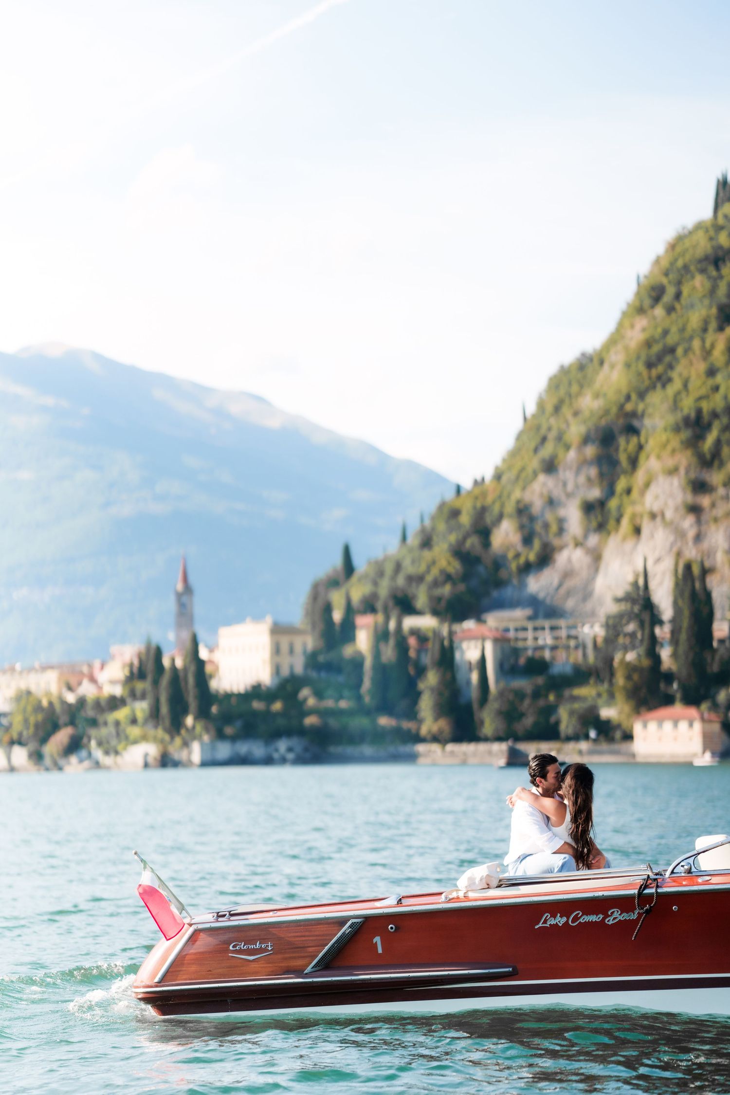 Couple embracing on a classic wooden speedboat on Lake Como, Italy, with mountain villages in the background.