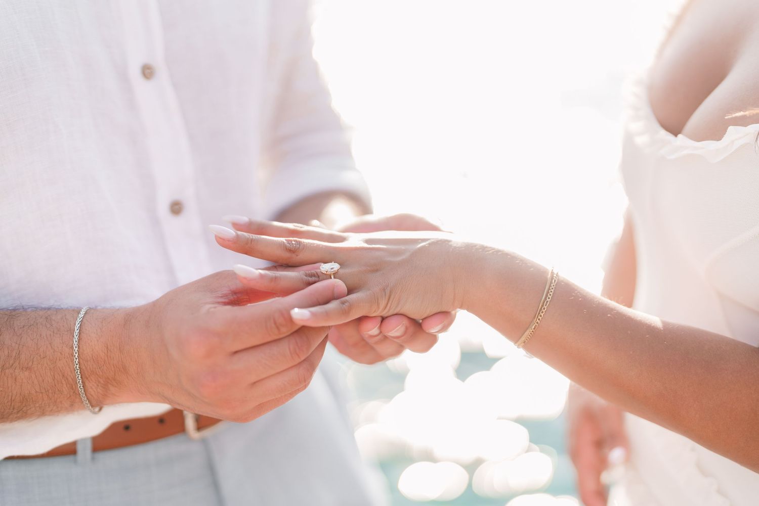 Groom placing a wedding ring on bride's finger during an outdoor beach ceremony in white attire.