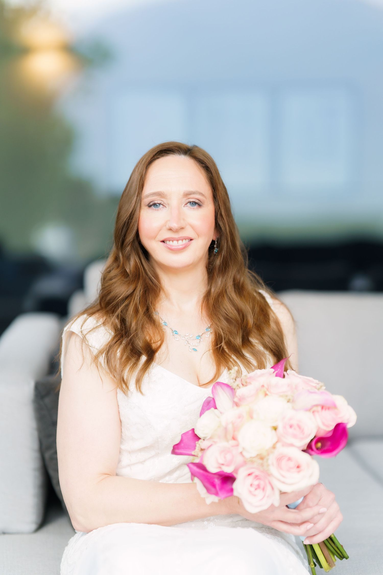 Smiling bride in white dress holding pink and white floral bouquet, seated outdoors with soft bokeh background.