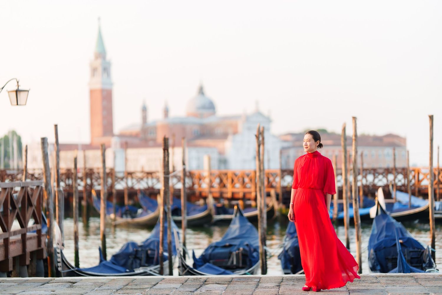 Woman in red gown poses by Venice gondolas with San Giorgio Maggiore church in soft morning light.