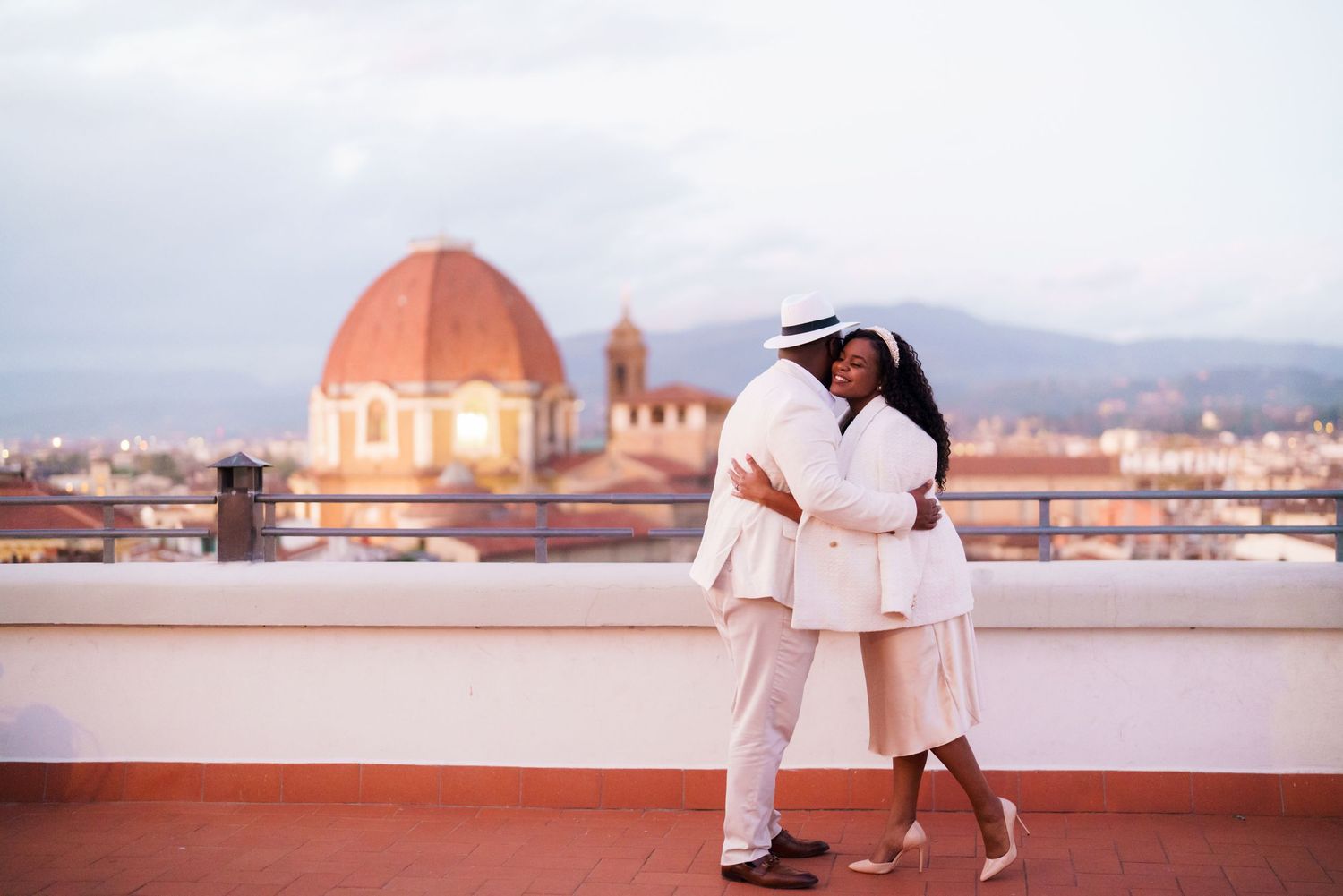 Couple in white outfits embracing on rooftop terrace with Florence Cathedral dome backdrop at dusk.
