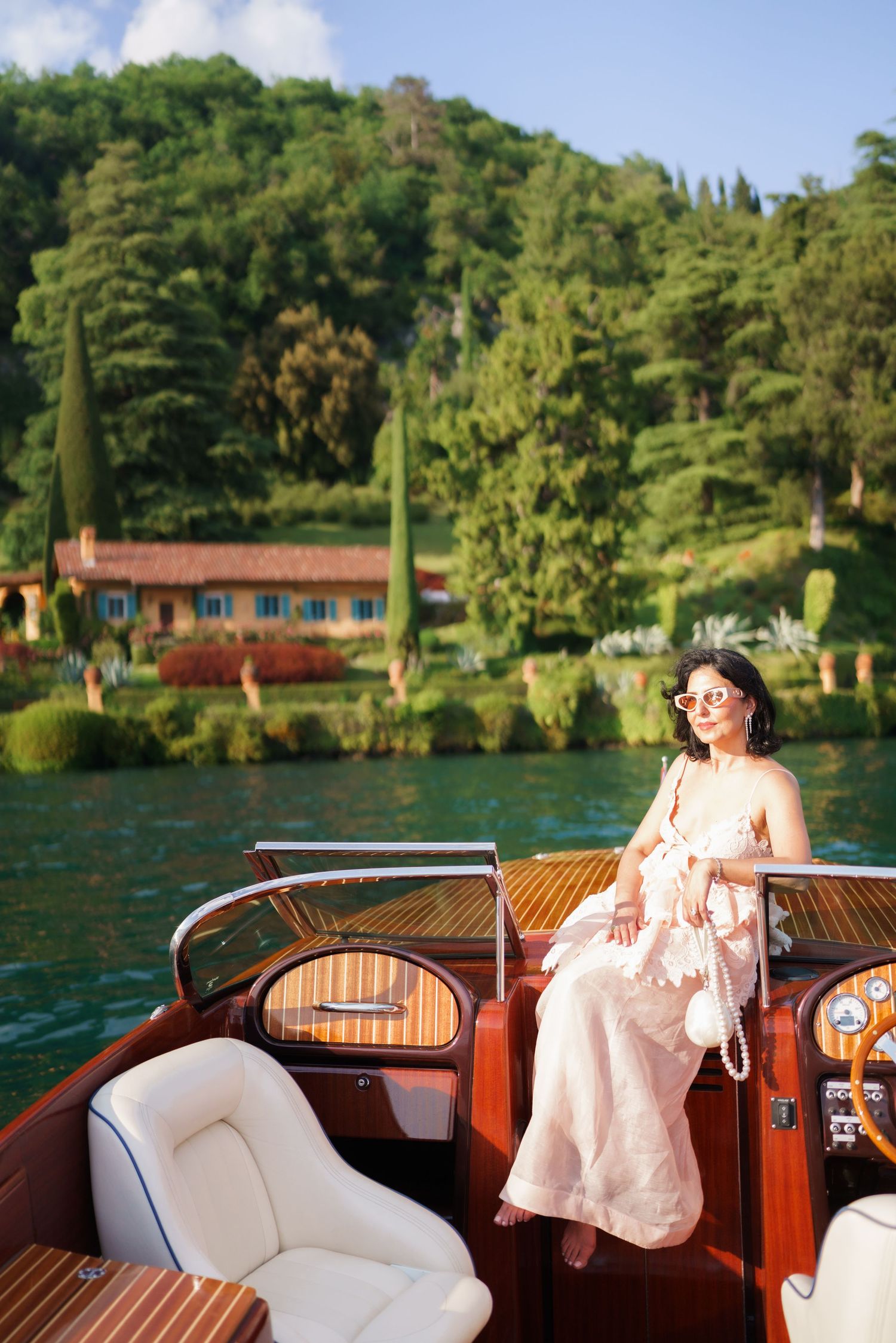 Woman in pink dress sitting on a luxury wooden boat on a lake with lush green hillside villa in background.