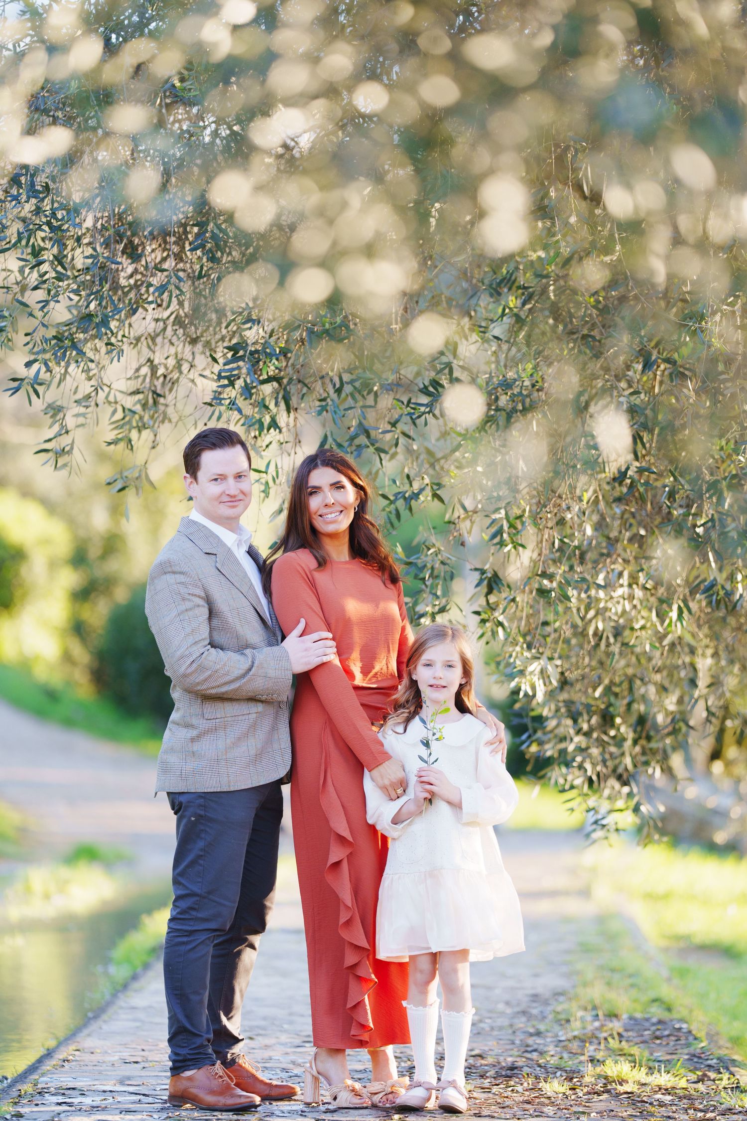 A happy family of three poses outdoors near a tree, with parents in elegant attire and daughter holding a flower.