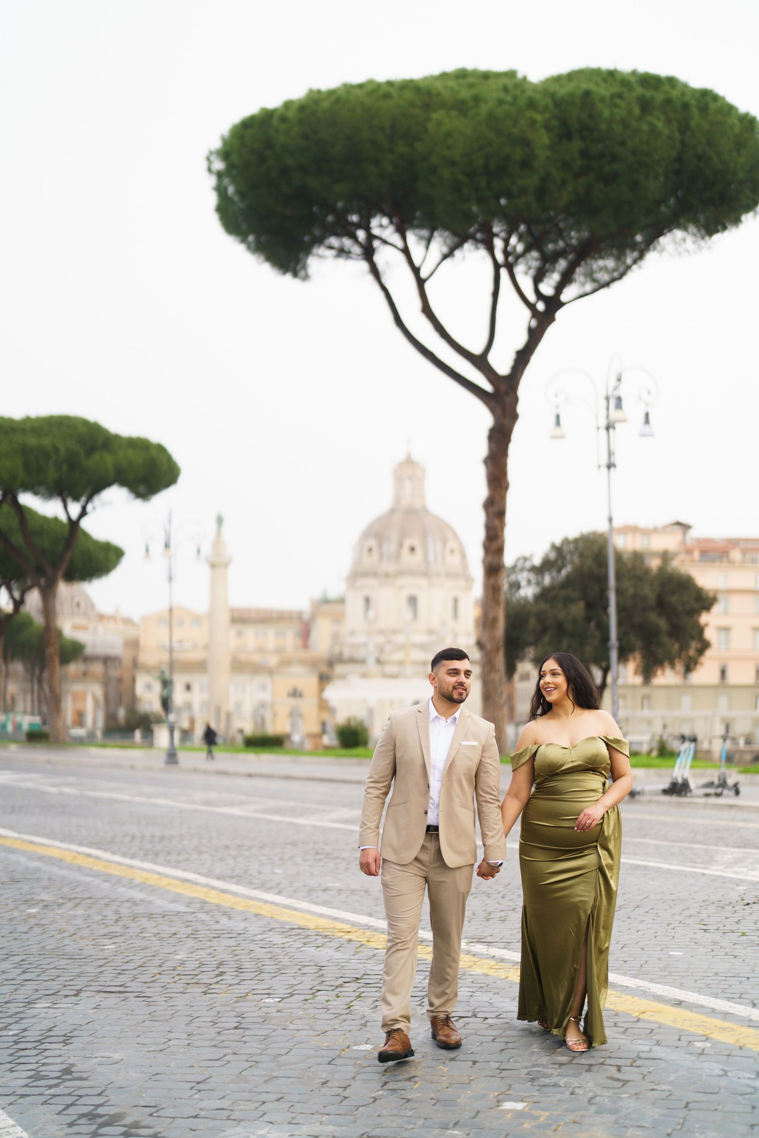 Couple holding hands walking on cobblestone street in Rome, Italy, with iconic umbrella pines and historic dome backdrop.