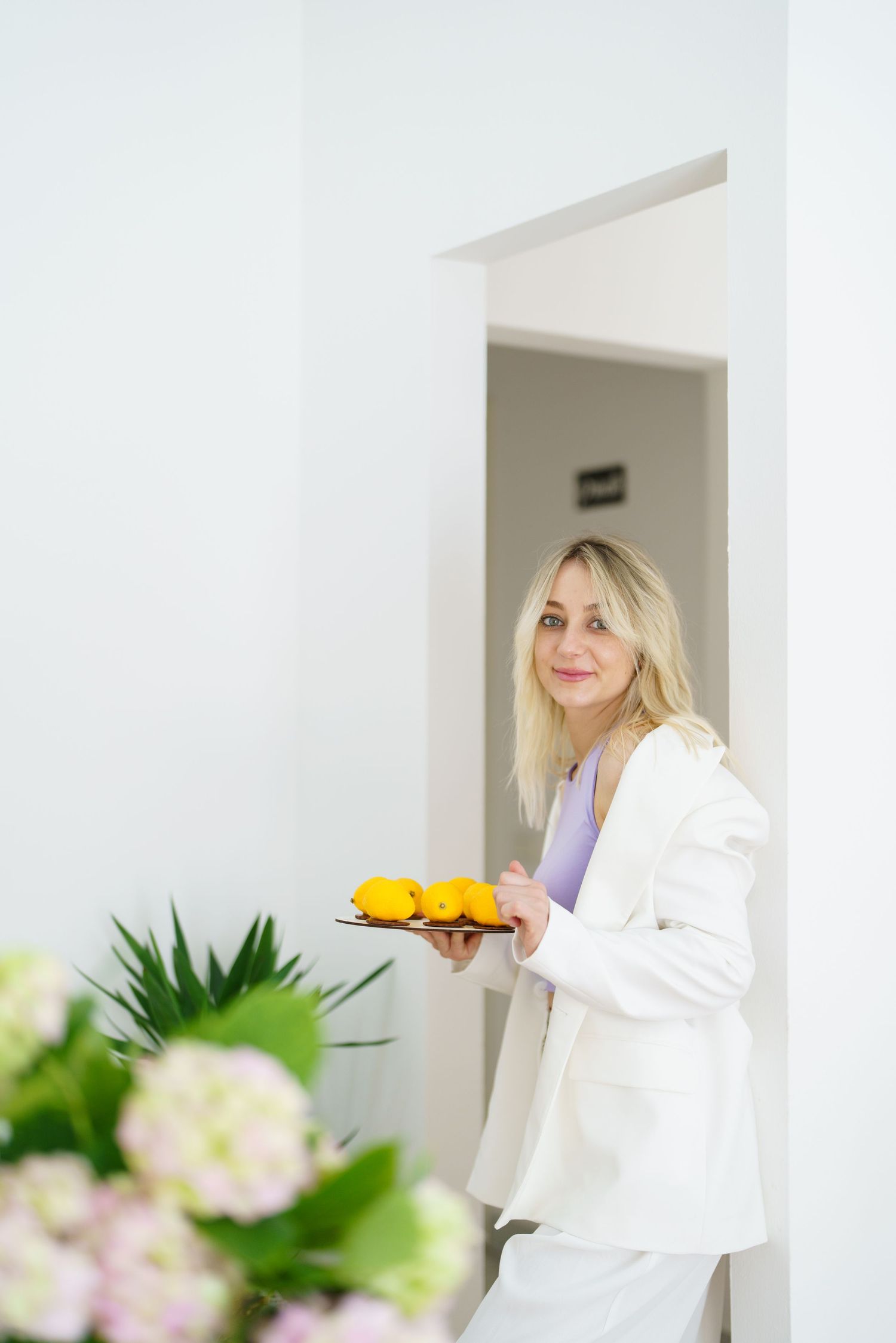 Blonde woman in white suit holding a tray of yellow lemons, standing in a bright white doorway with flowers in foreground.