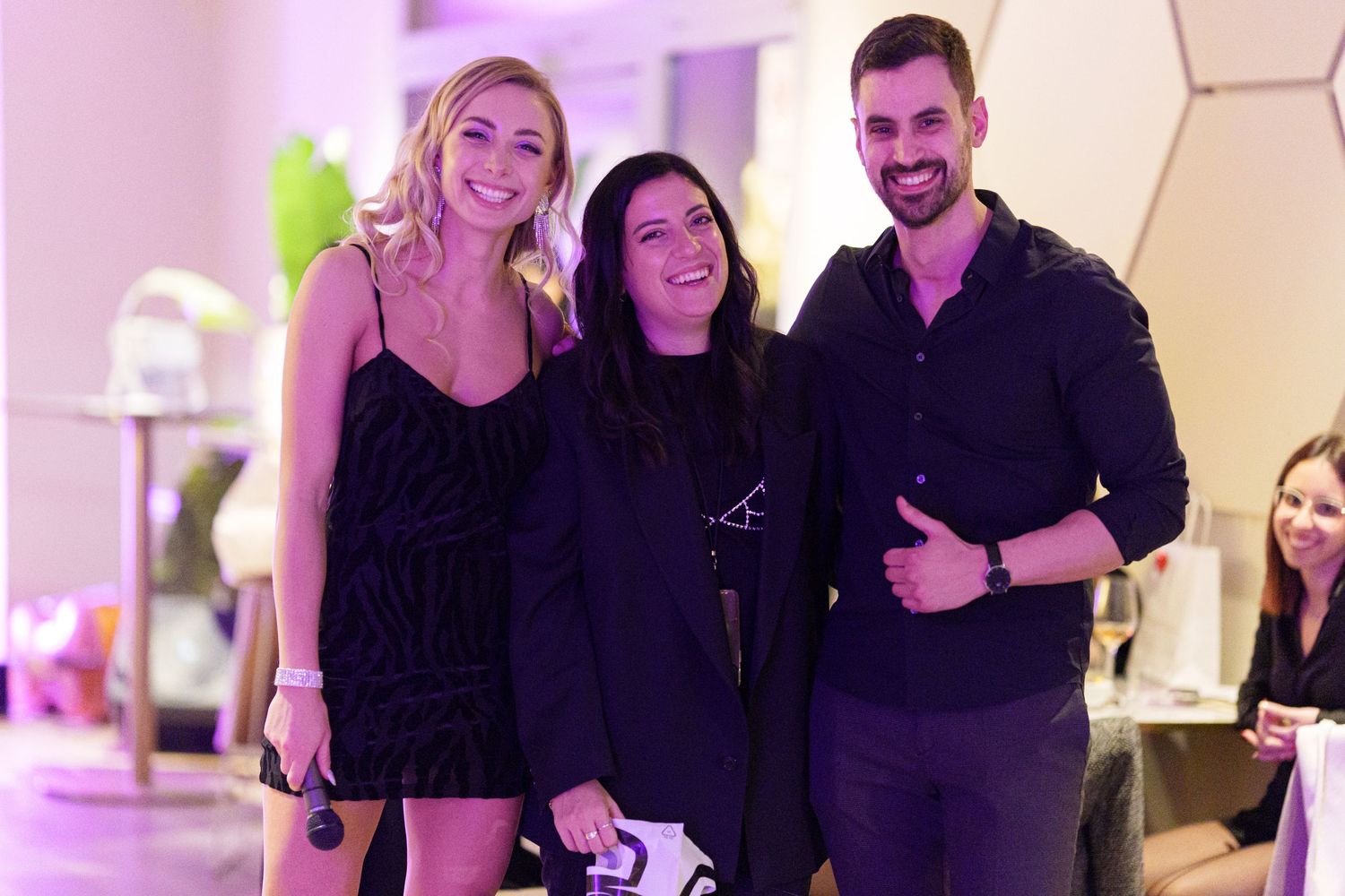 Three smiling people posing together at an indoor event, two women and a man giving a thumbs up.