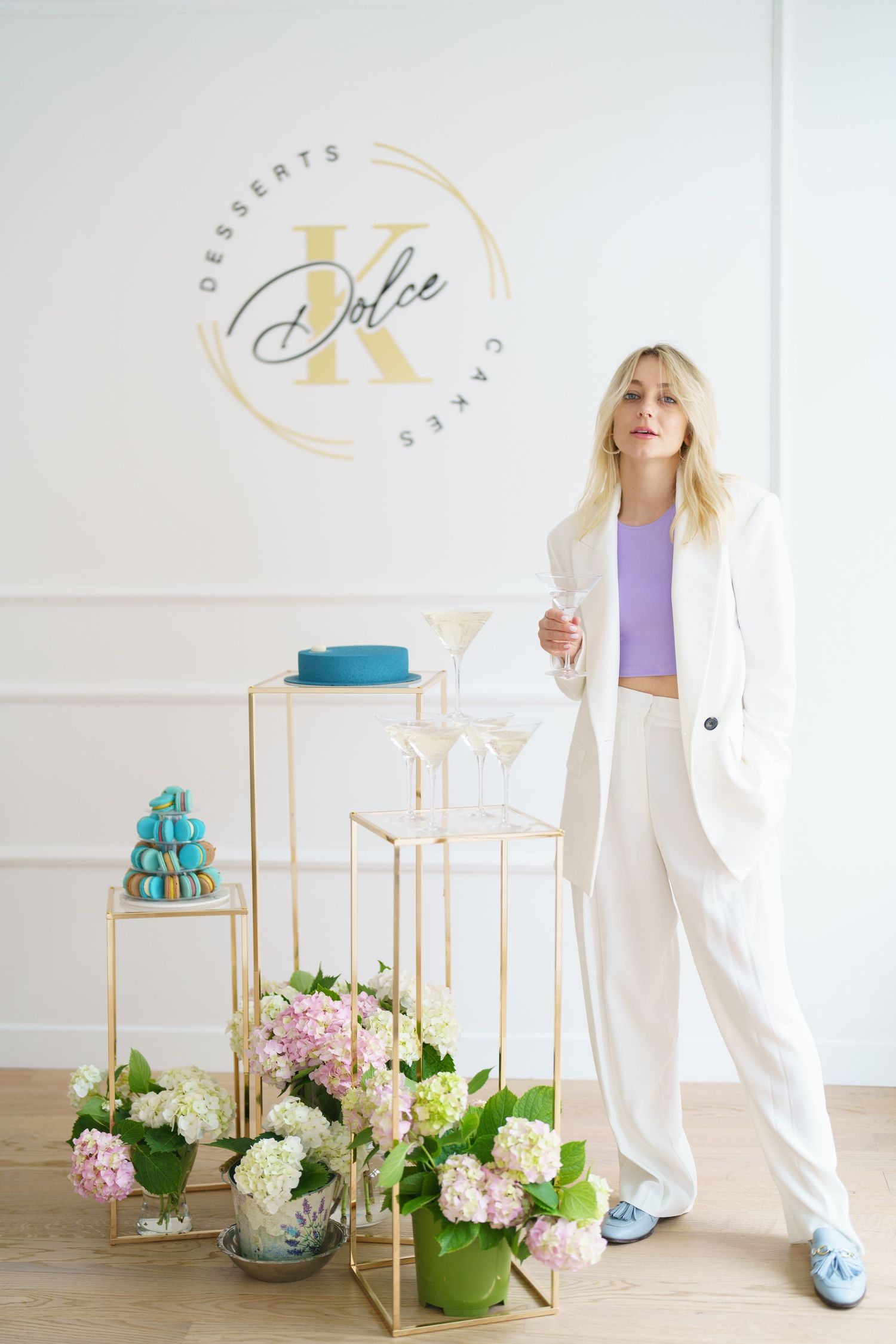 Blonde woman in white suit poses beside gold stands with macarons, blue cake, and hydrangeas at Dolce K Desserts.