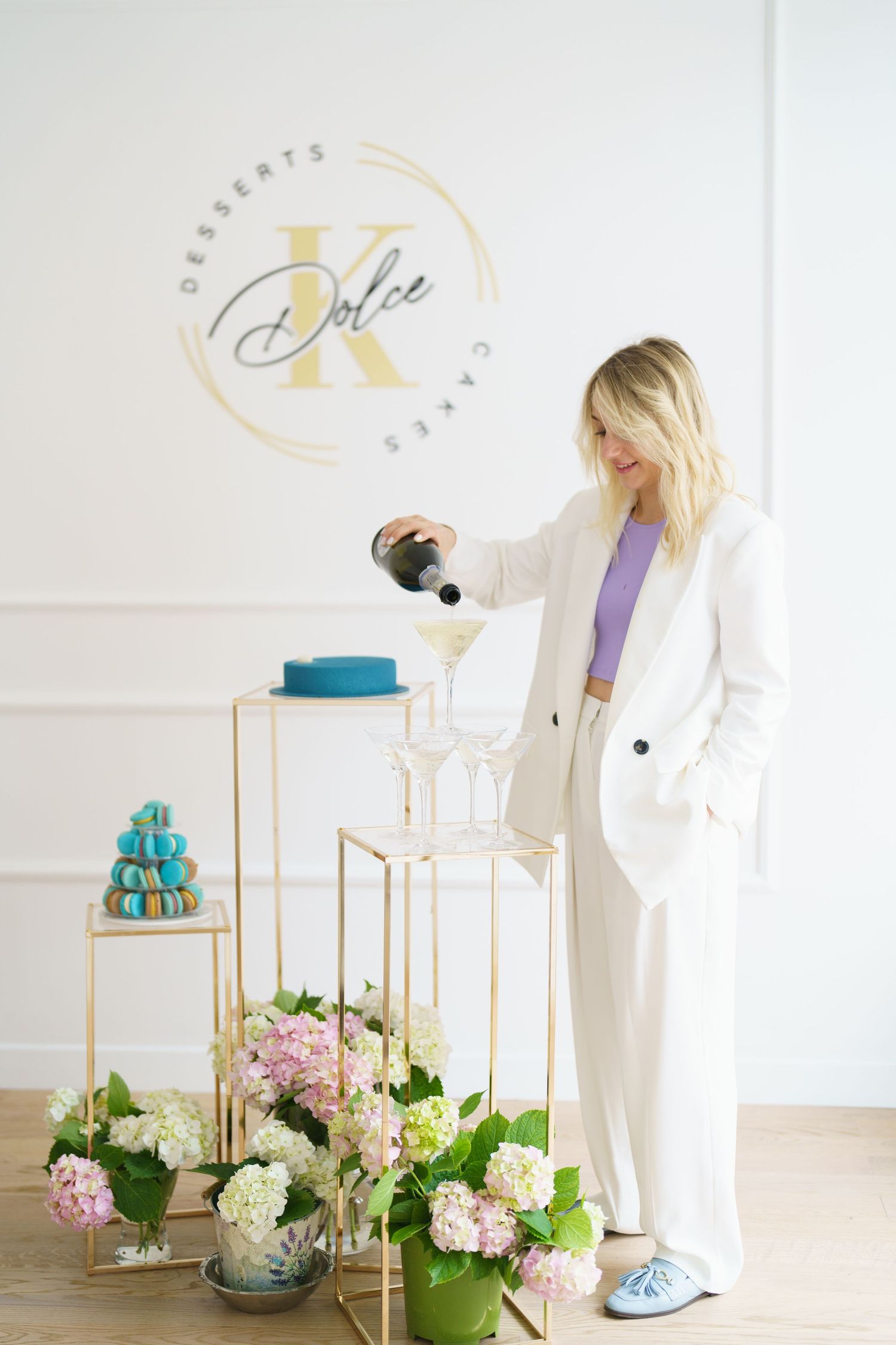 Woman in white suit pouring champagne tower at Dolce K Desserts Cakes shop with floral arrangements.