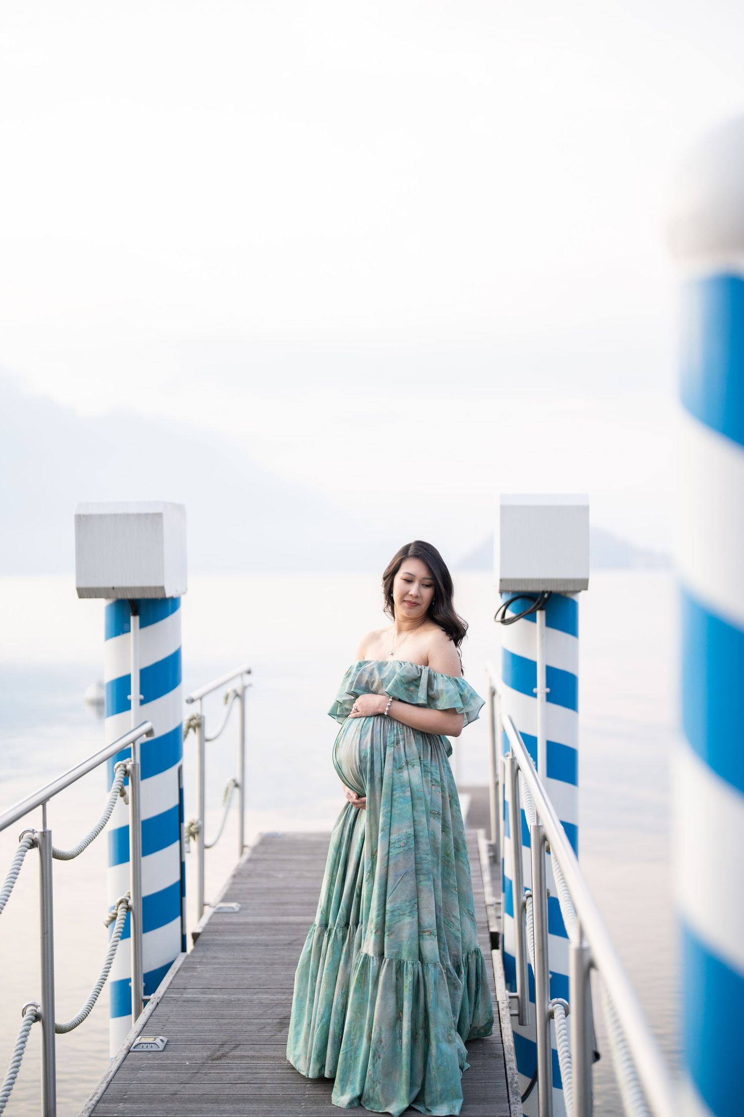 Pregnant woman in green off-shoulder maxi dress standing on a lakeside dock with blue and white pillars.