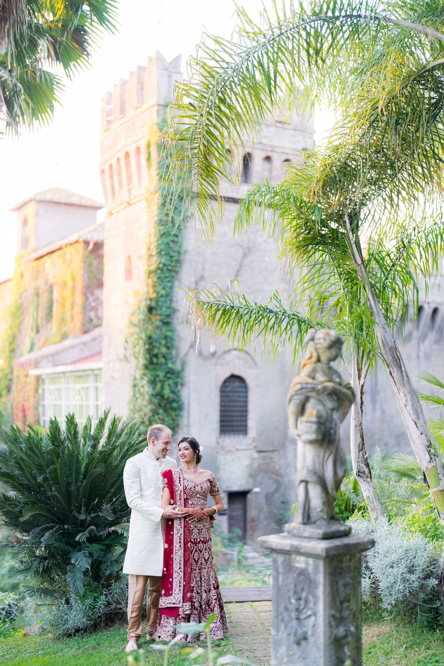 A romantic couple poses in front of a castle-like building with palm trees and stone statues.