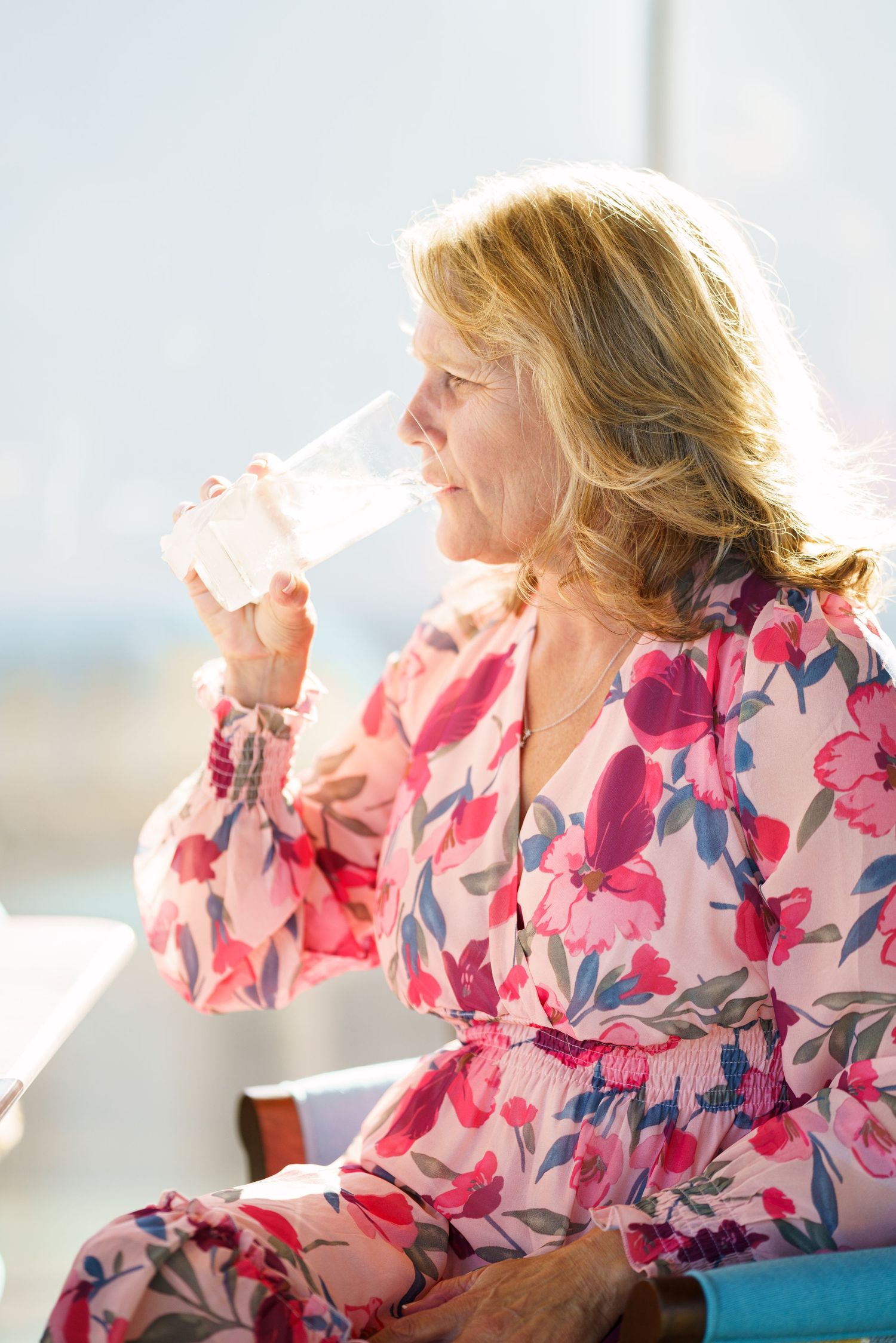Person in floral pink dress sipping from glass near window.