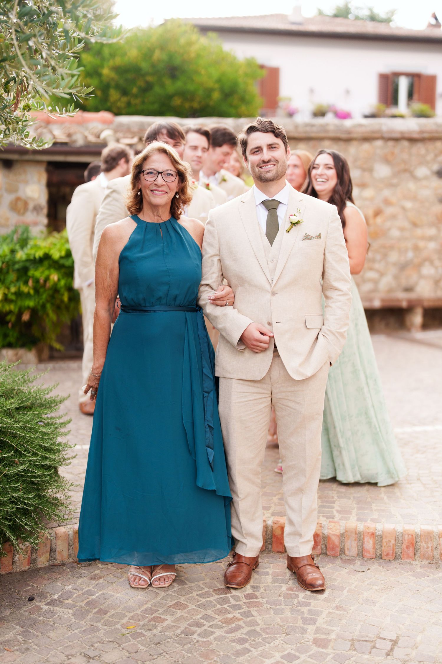 Wedding party members in cream suits and teal dresses pose on stone walkway.