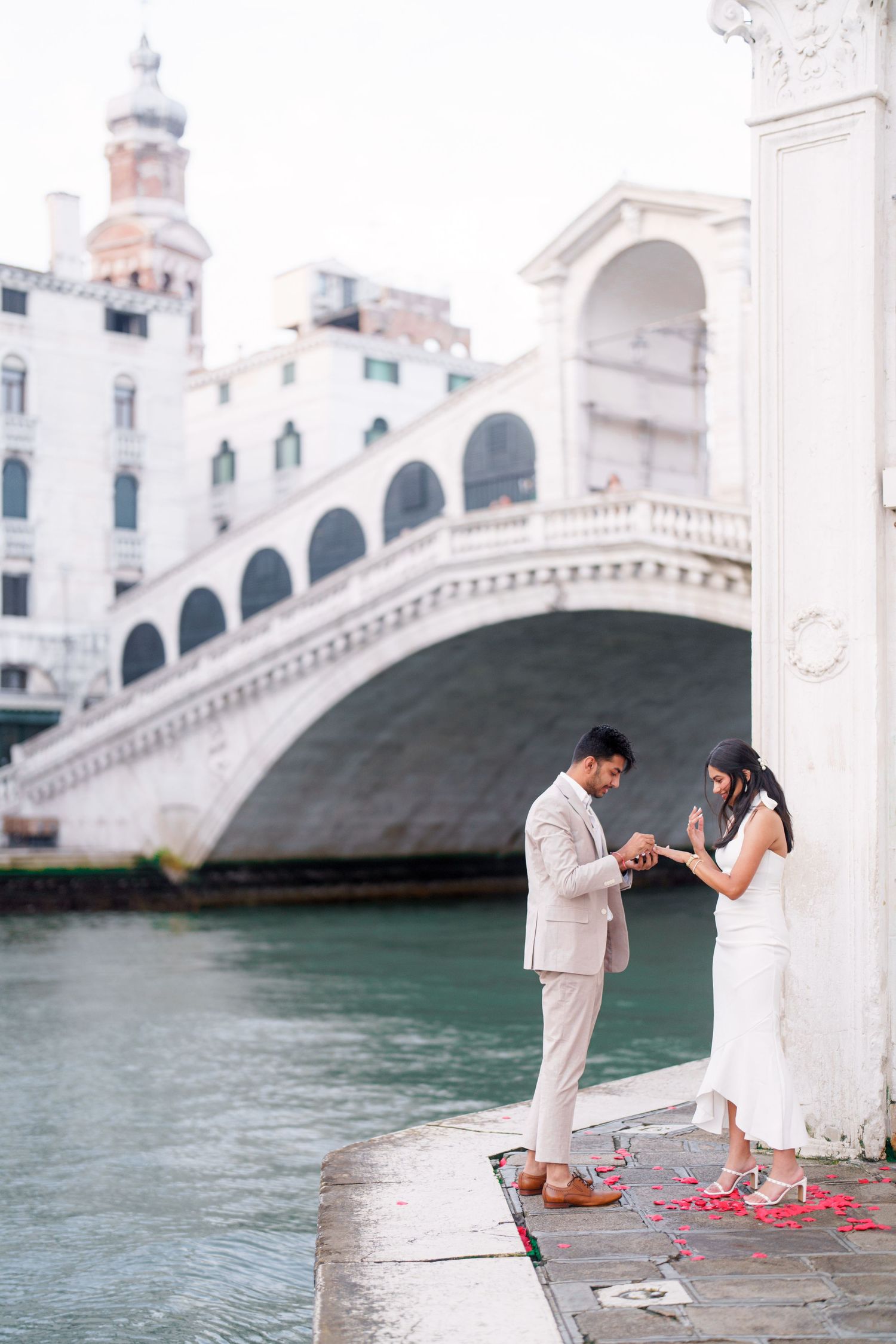 A romantic proposal scene unfolds on a stone bridge over turquoise waters in Venice, Italy.