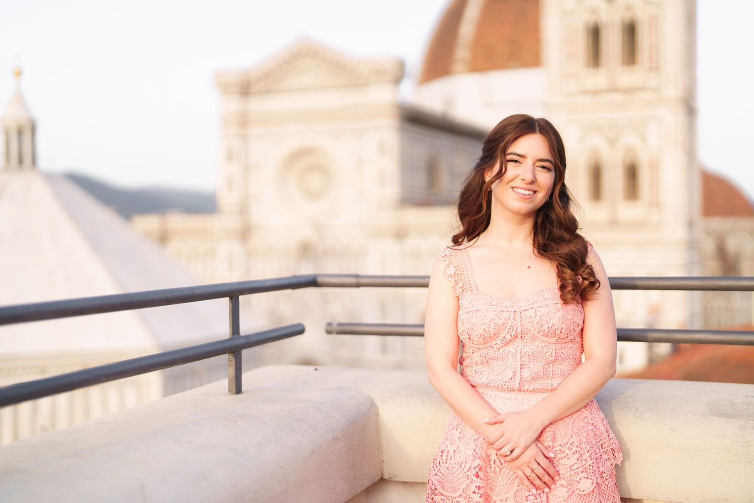 Woman in peach-colored dress poses on balcony with European architecture in background during golden hour.