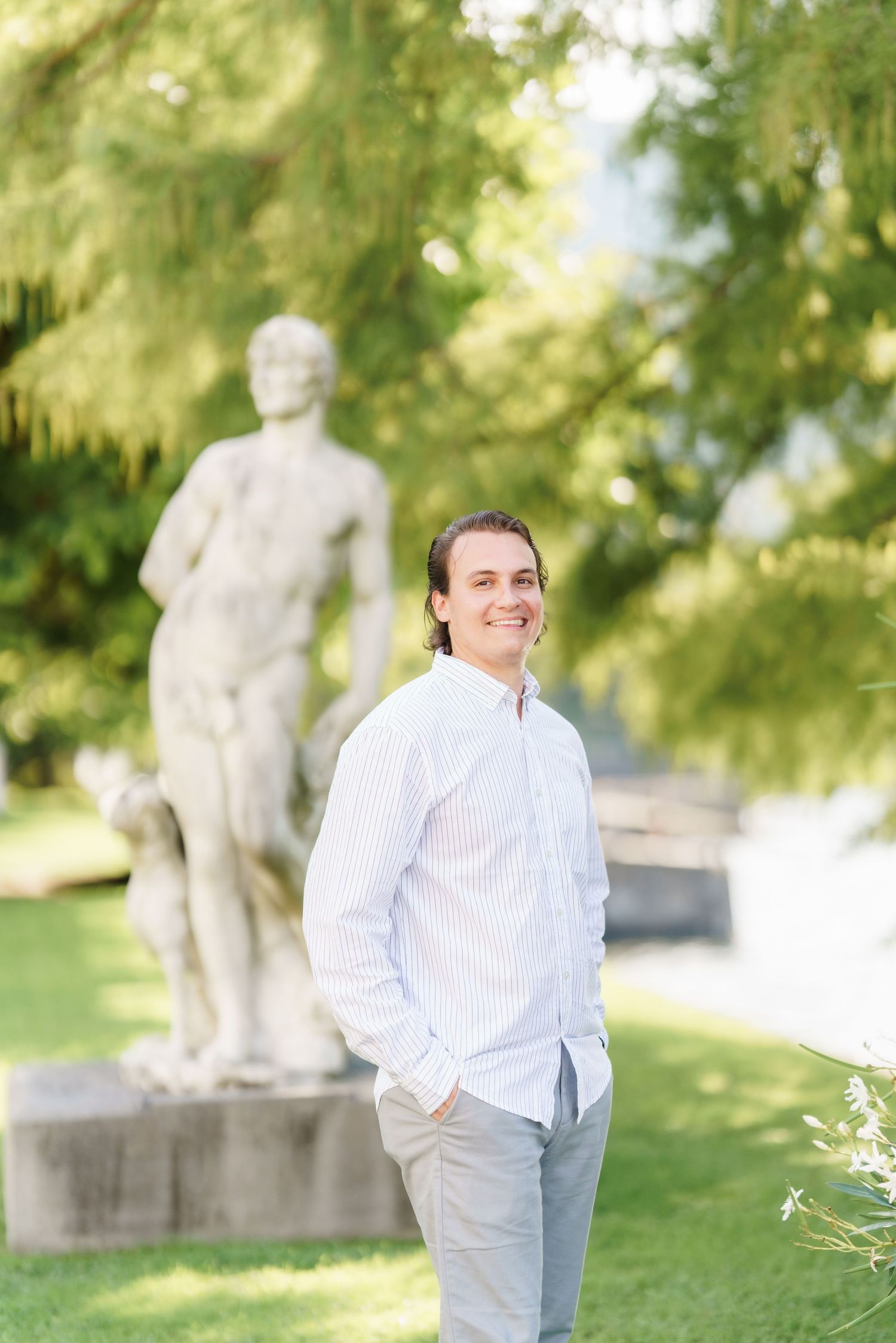 A person in a white button-down shirt stands in a sunny garden setting with stone statues visible in the background.