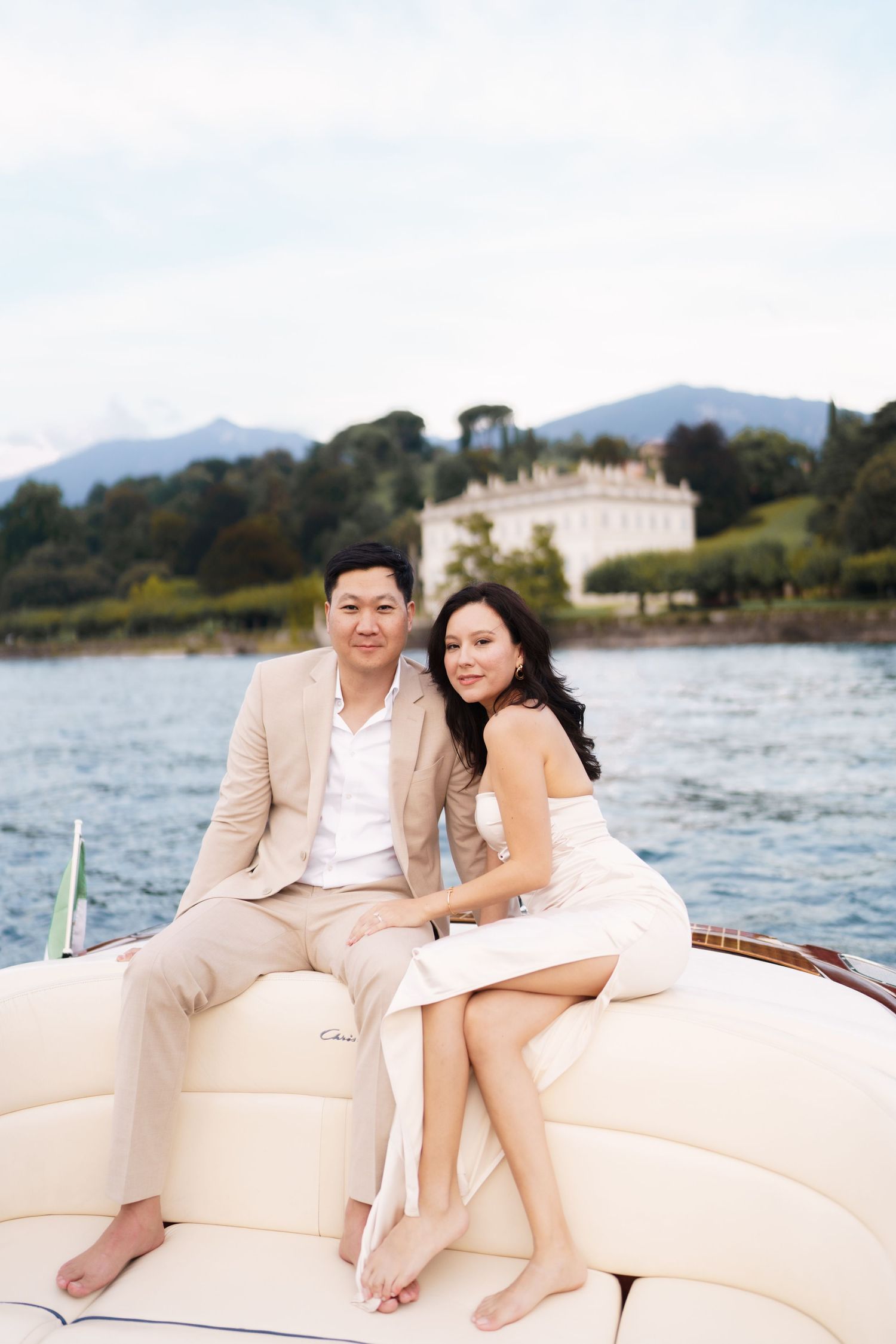 A couple in light colored outfits sits together on a boat with mountains and a villa visible in the background.