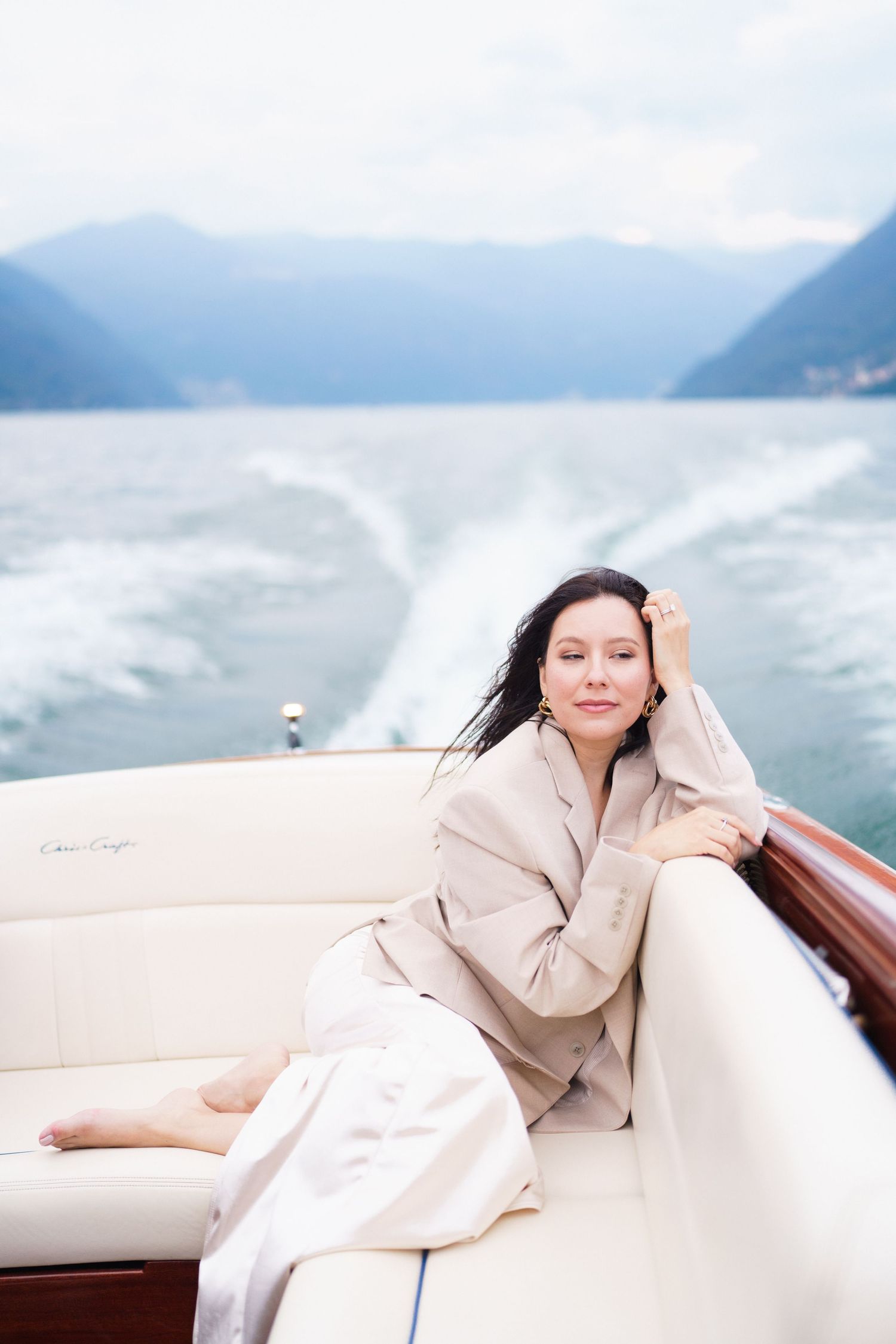 A person in cream colored outfit relaxes on a luxury boat with misty mountains and water in the background.