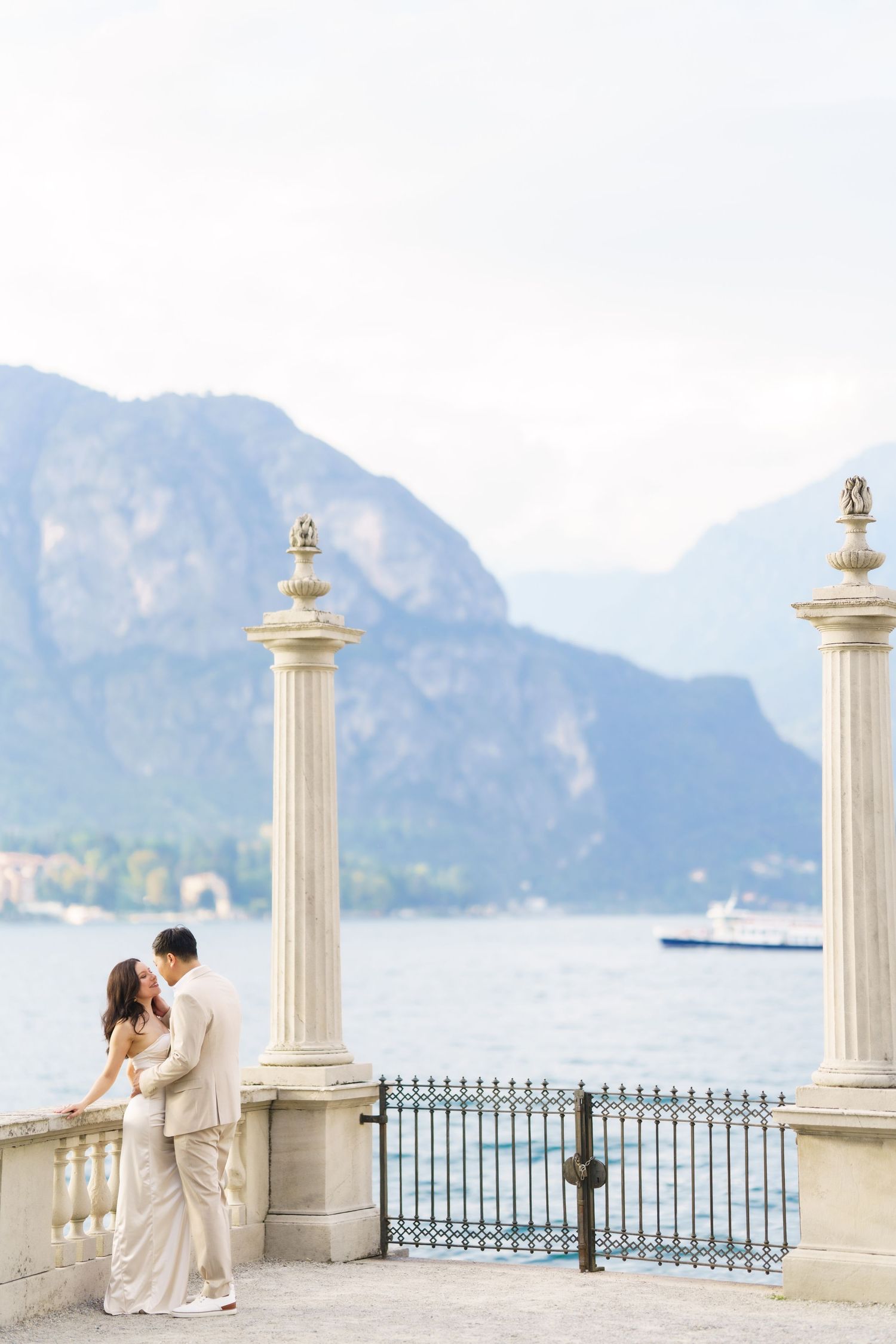 A couple embraces on an elegant lakeside terrace with ornate white columns and mountain views at Lake Como, Italy.