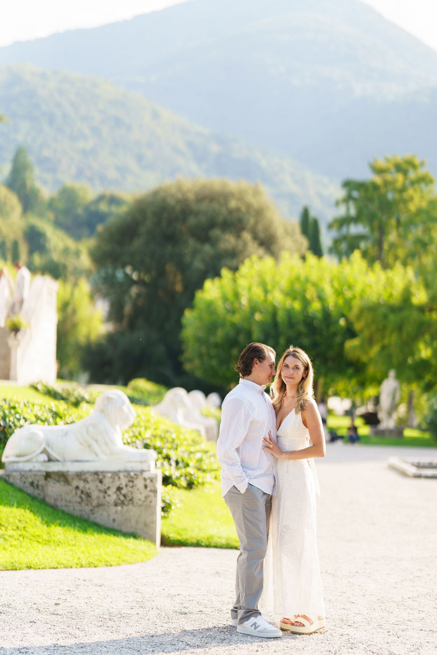 A couple in casual white outfits embrace on a garden path with mountains and lush greenery in the background.