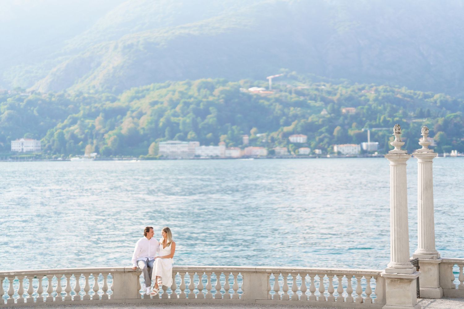 A couple stands on an elegant white stone balcony overlooking a scenic mountain lake on a sunny day.