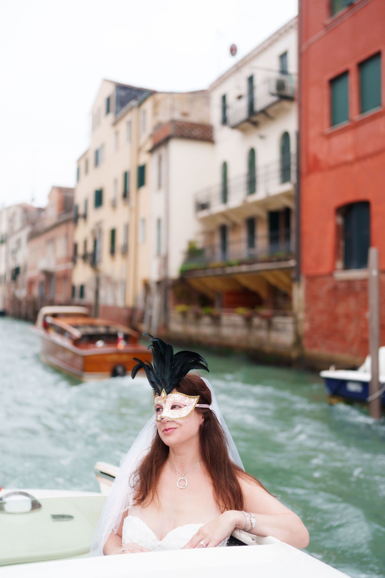 A masked figure in a Venetian carnival mask sits in a boat on a canal in Venice with colorful buildings in the background.
