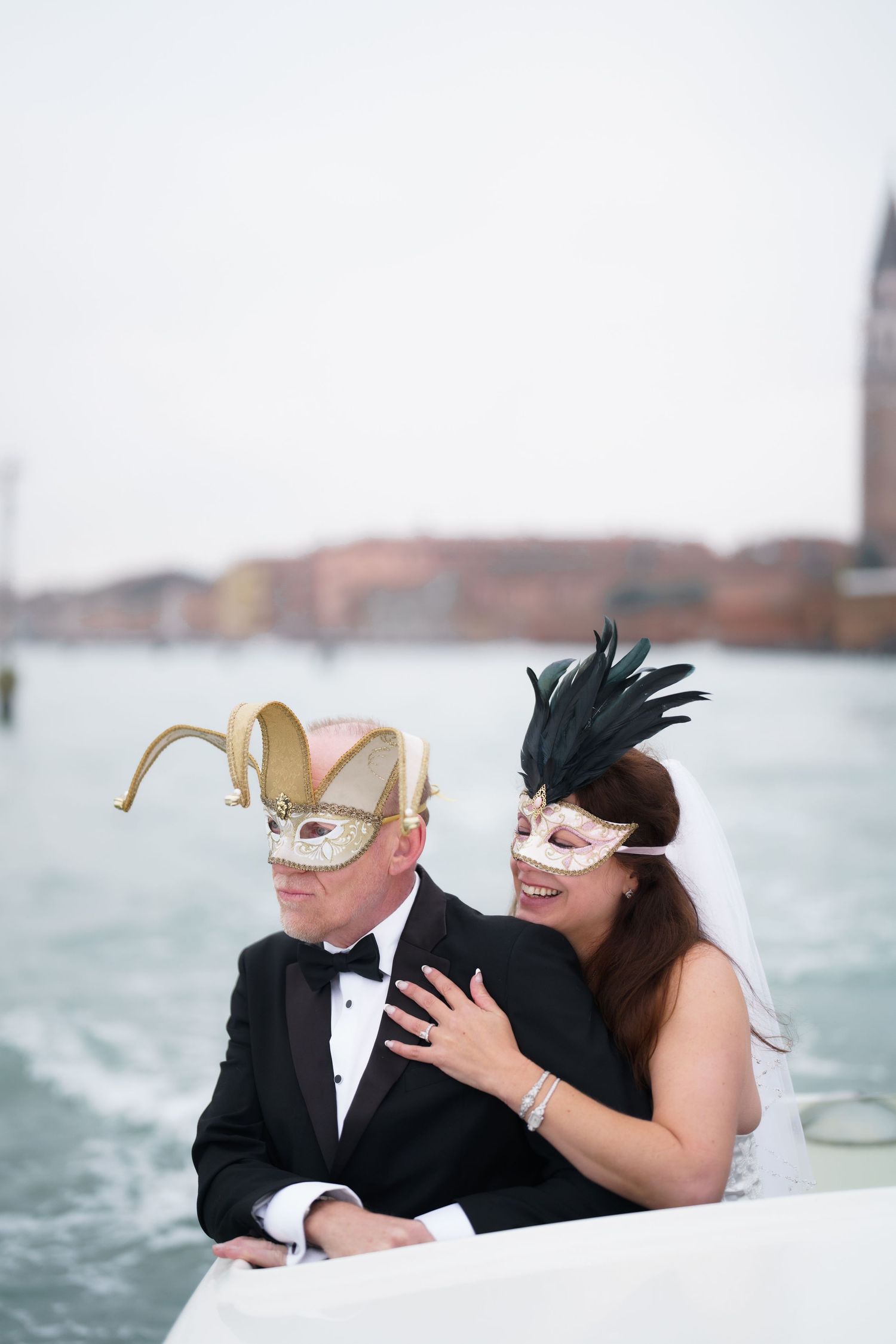 A couple in formal attire and masquerade masks share an intimate moment by the water in Venice, Italy.