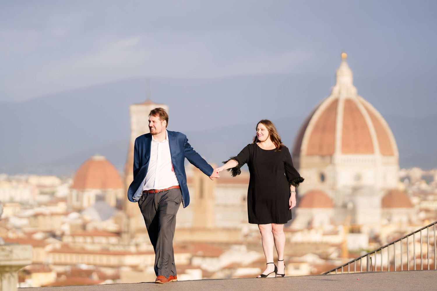 A couple holds hands while walking along a scenic overlook with Florence's Duomo cathedral dome in the background.