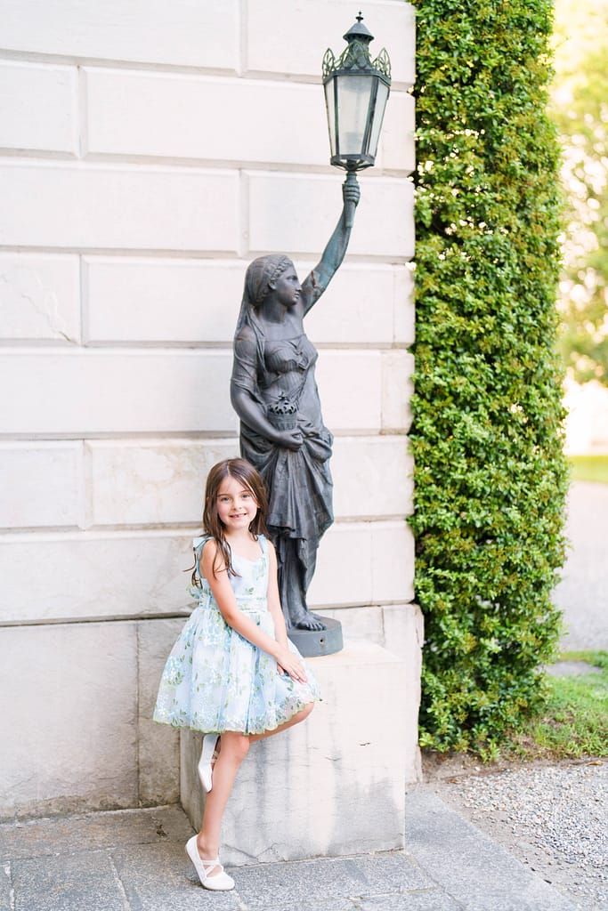 A decorative statue holds a lamp post on a white building wall while a young person in a blue dress poses beside it.
