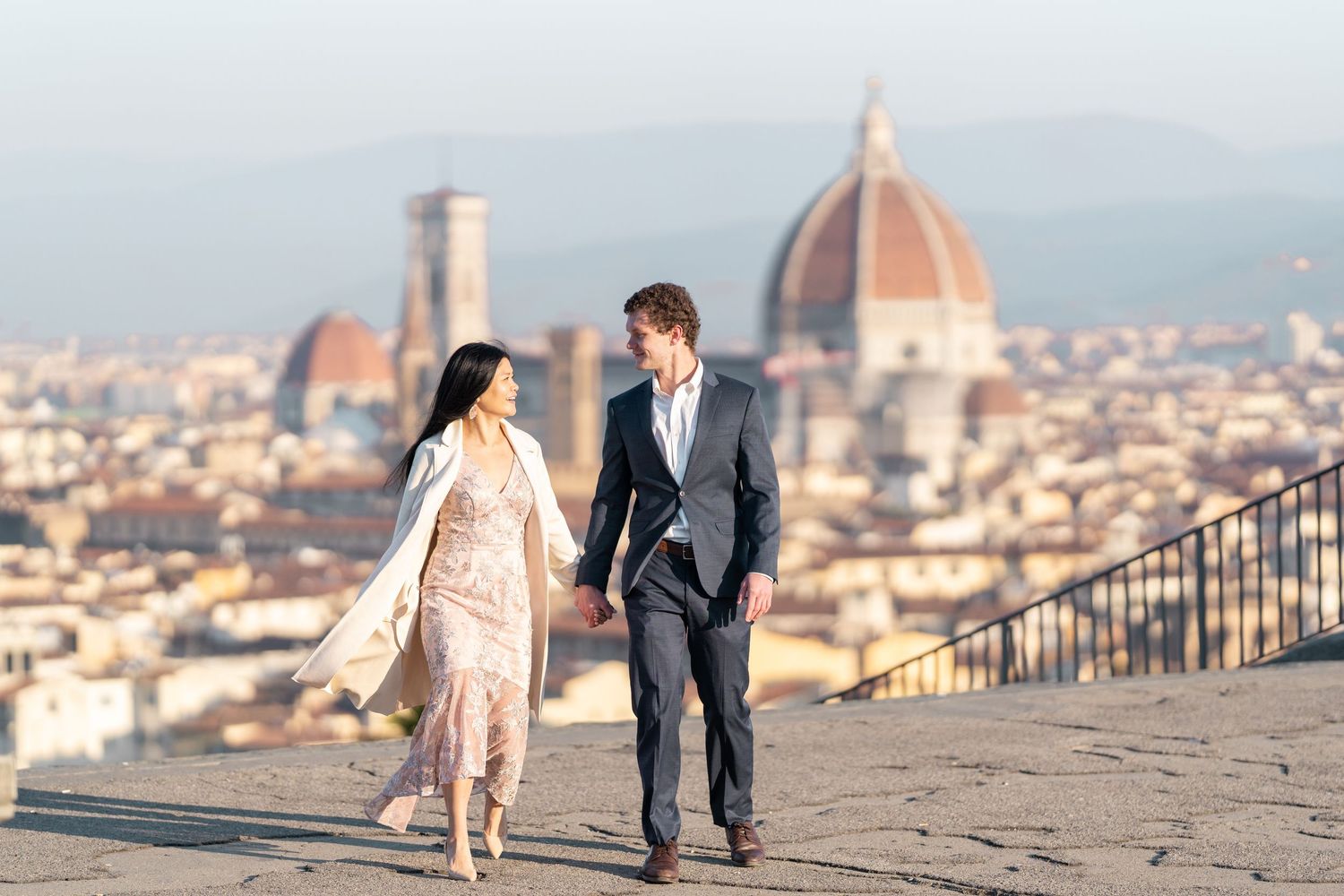 A couple walks along a scenic overlook with the Florence Cathedral and cityscape visible in the background.
