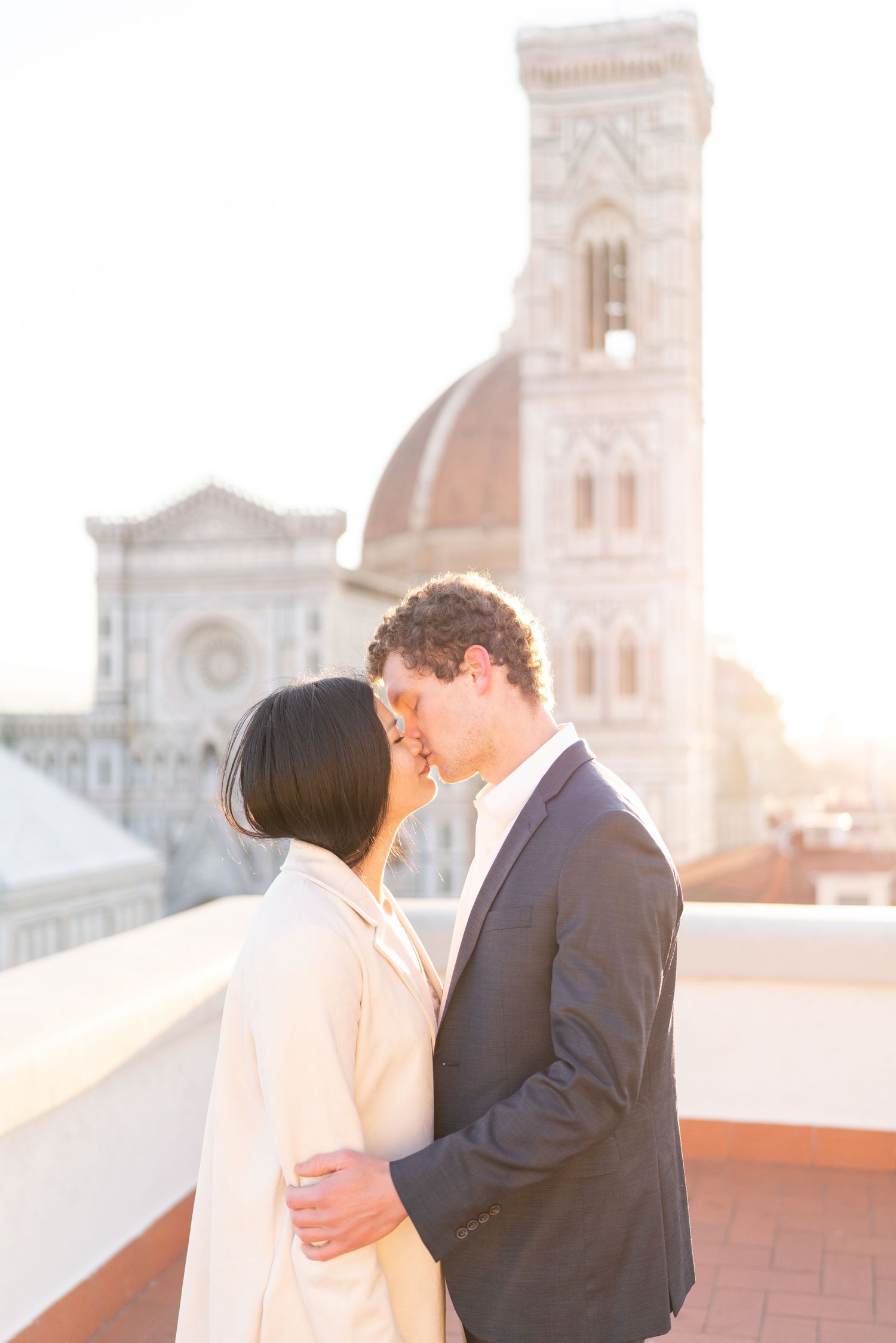 A couple shares a romantic kiss on a rooftop terrace at sunset with a beautiful church dome visible in the background.