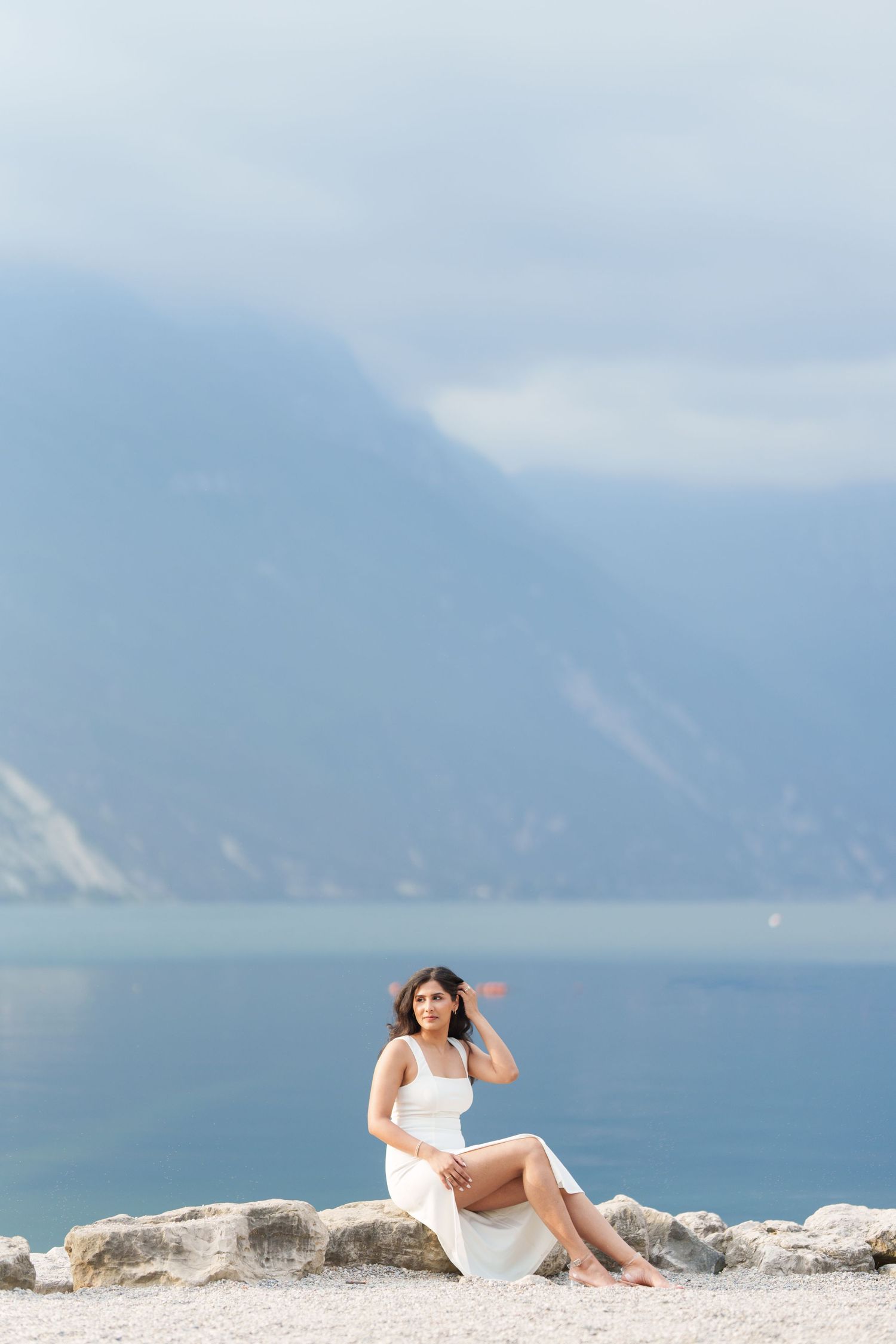A person in a white dress reclines on coastal rocks overlooking a misty ocean and mountain landscape.