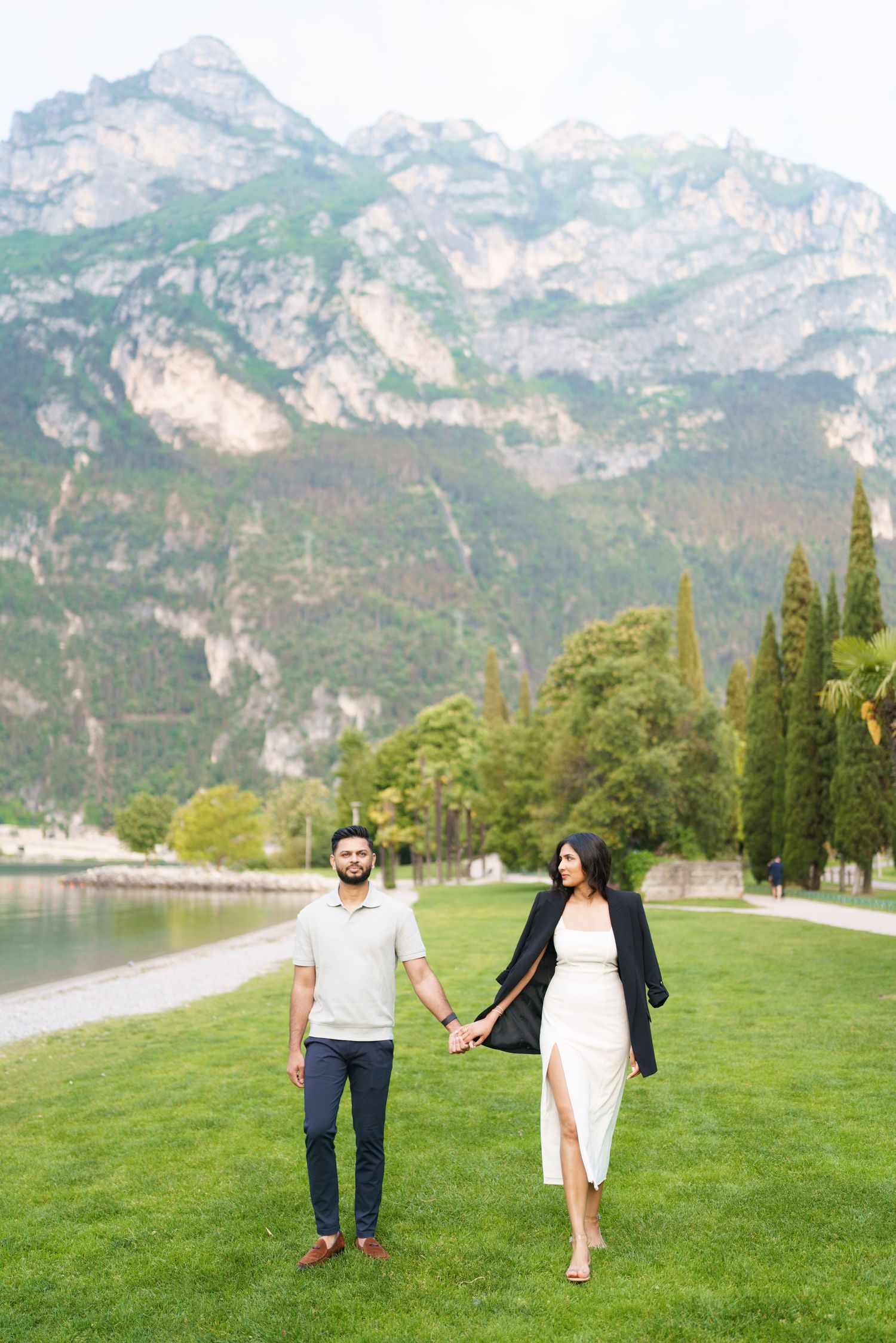 A couple walks hand in hand along a lakeside path with dramatic mountain peaks and cypress trees in the background.