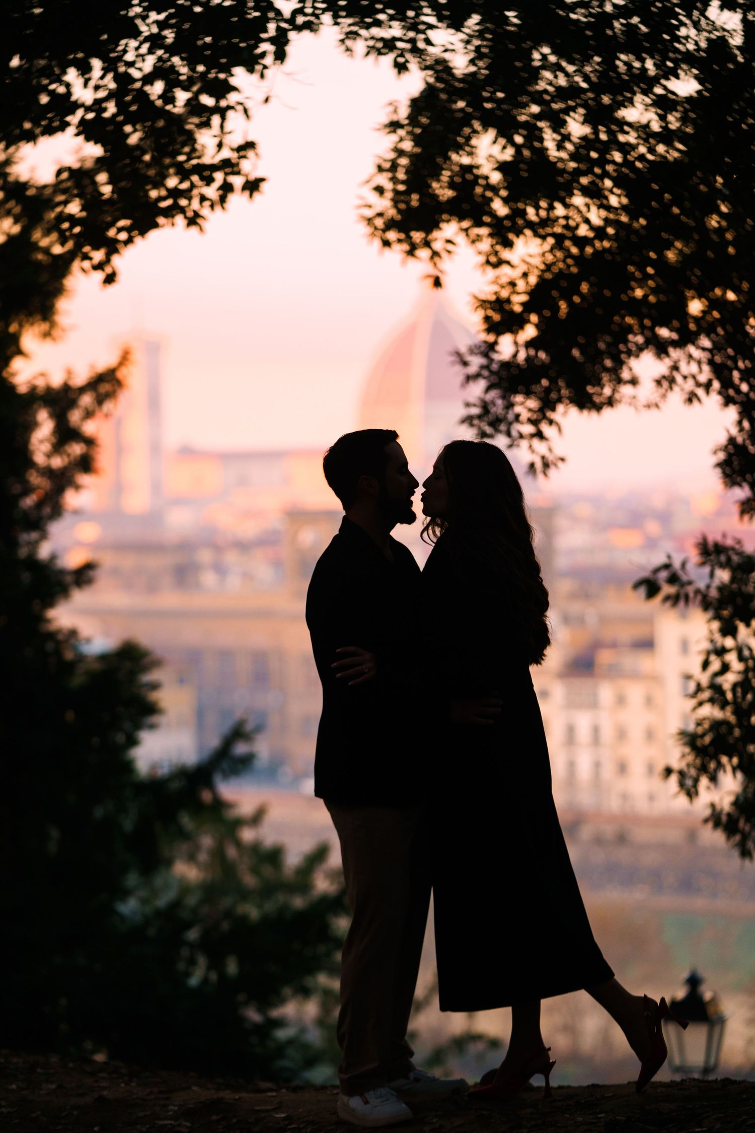 Silhouettes of a romantic couple kissing at sunset with trees and cityscape in the background.